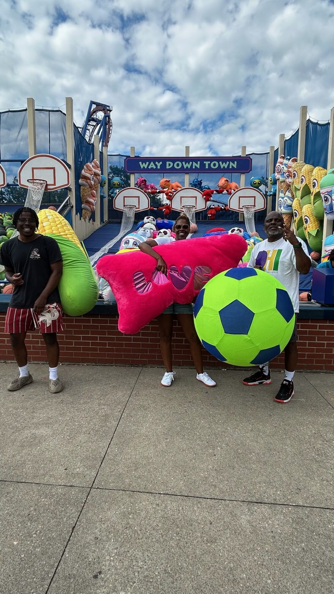 People holding inflatable toys in front of a colorful amusement park with a sign reading 'Way Down Town'.