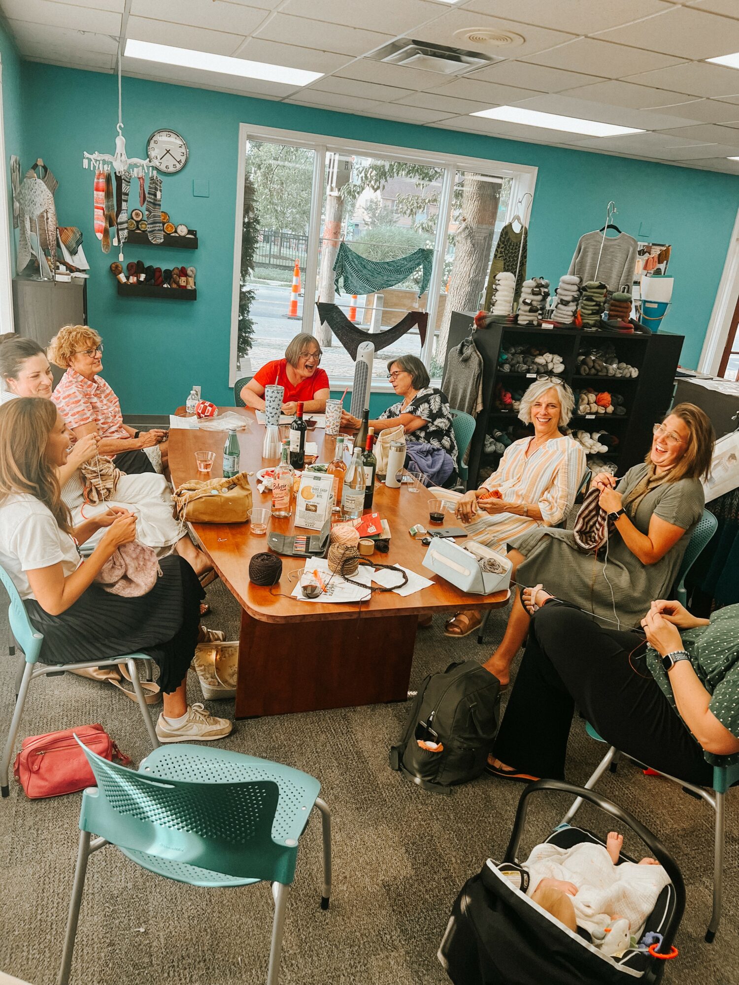 Group of women sitting around a table in a room with teal walls, large window, and shelves with various items.