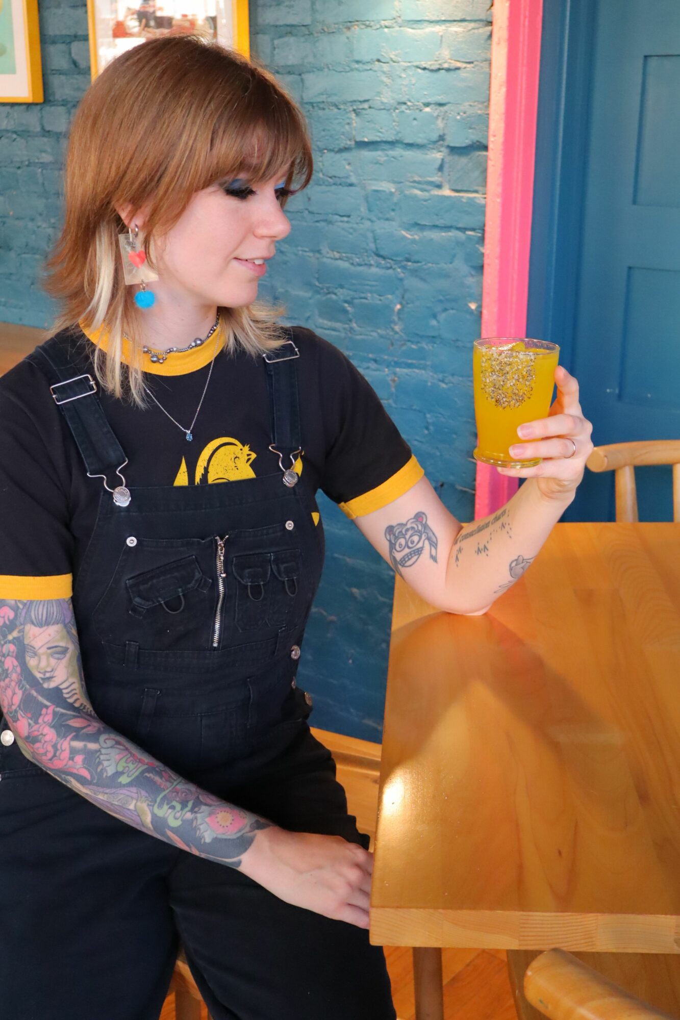 Woman with tattoos and earrings holding a yellow drink in a colorful room.