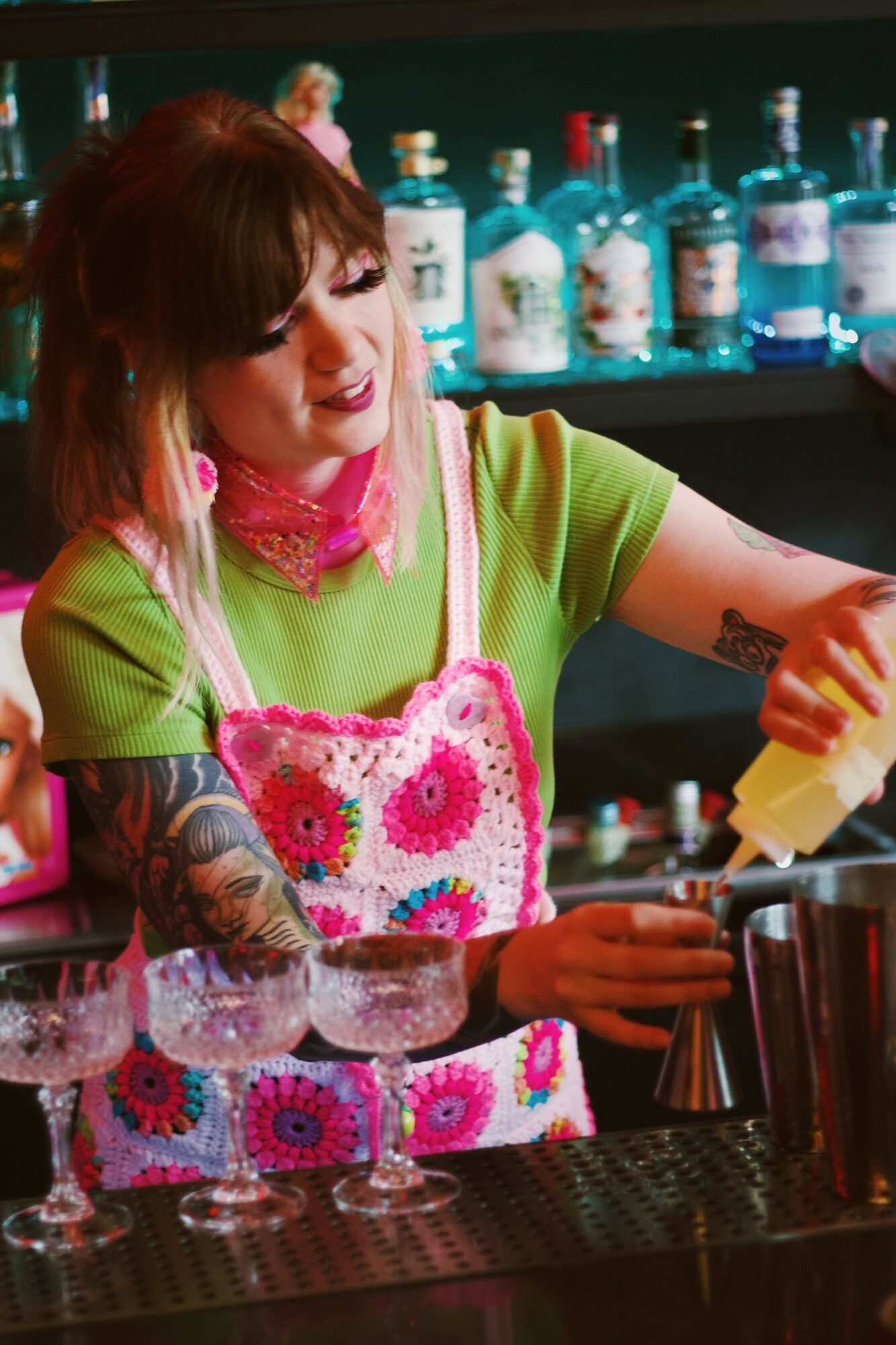 Person with pink hair and tattoos pouring a drink at a bar, wearing a green shirt and pink apron.