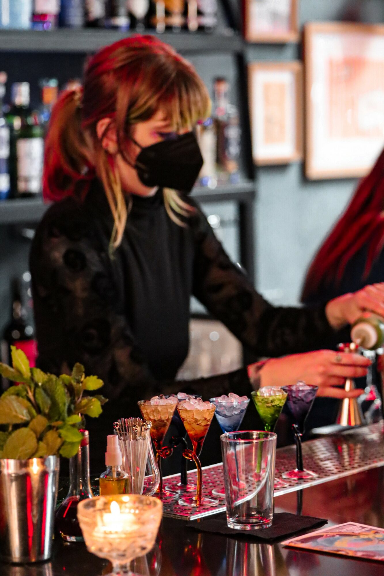 Bartender wearing a black mask and black outfit prepares drinks at a bar with colorful cocktails and plants.