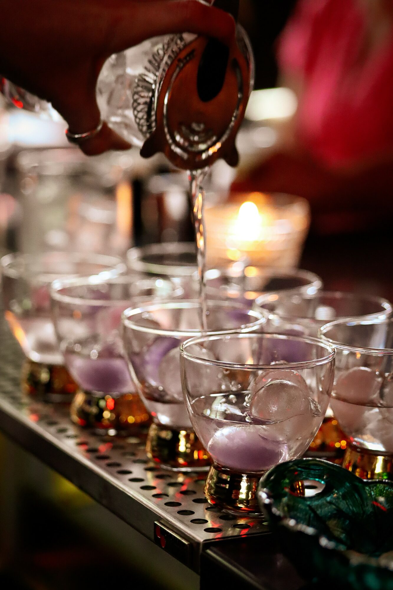 Person pouring liquid into glasses with purple liquid, glasses arranged on a metallic surface, warm background lighting.