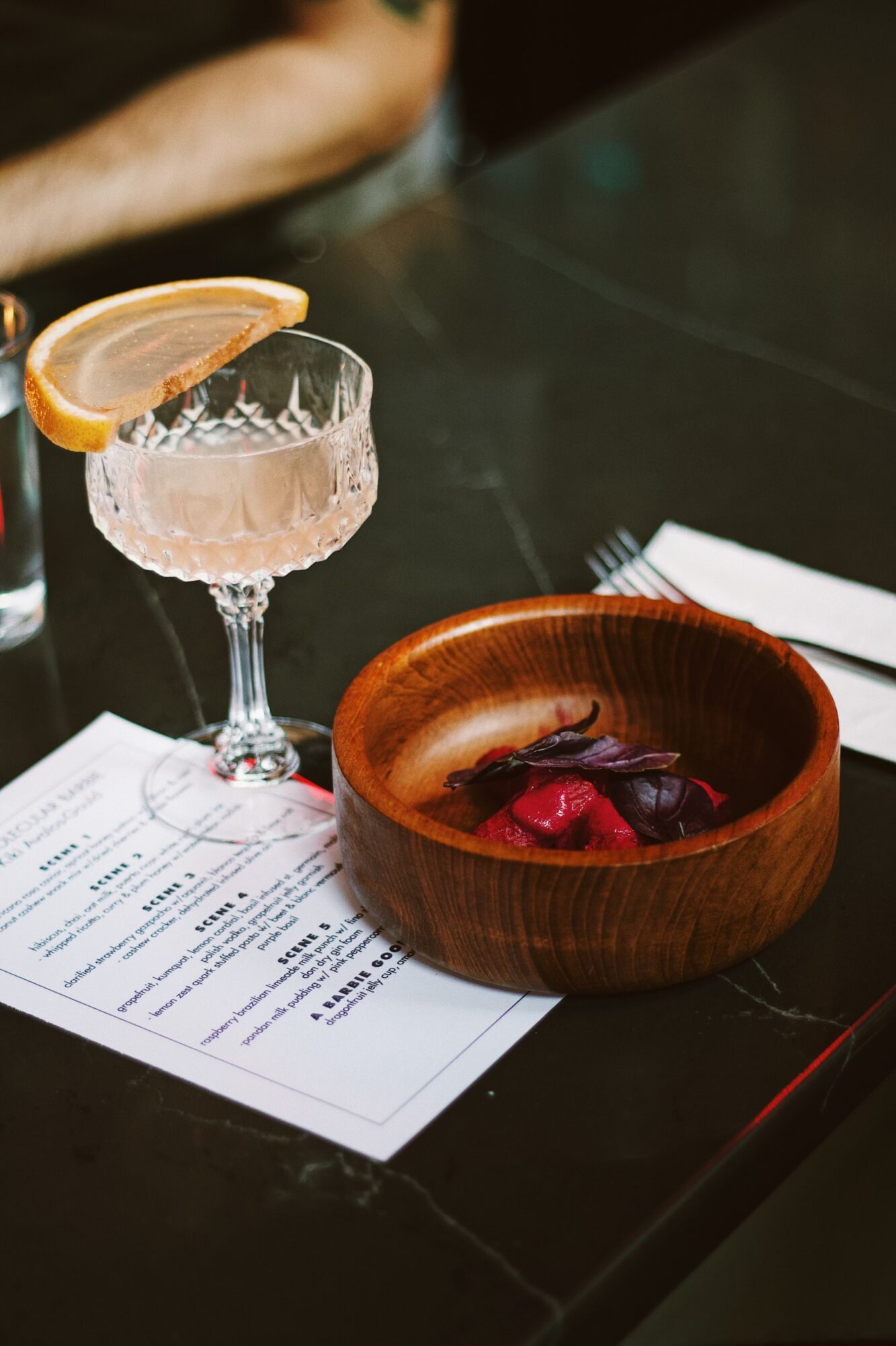 Cocktail glass with pink drink and lemon slice, wooden bowl with food, paper menu, dark table surface.