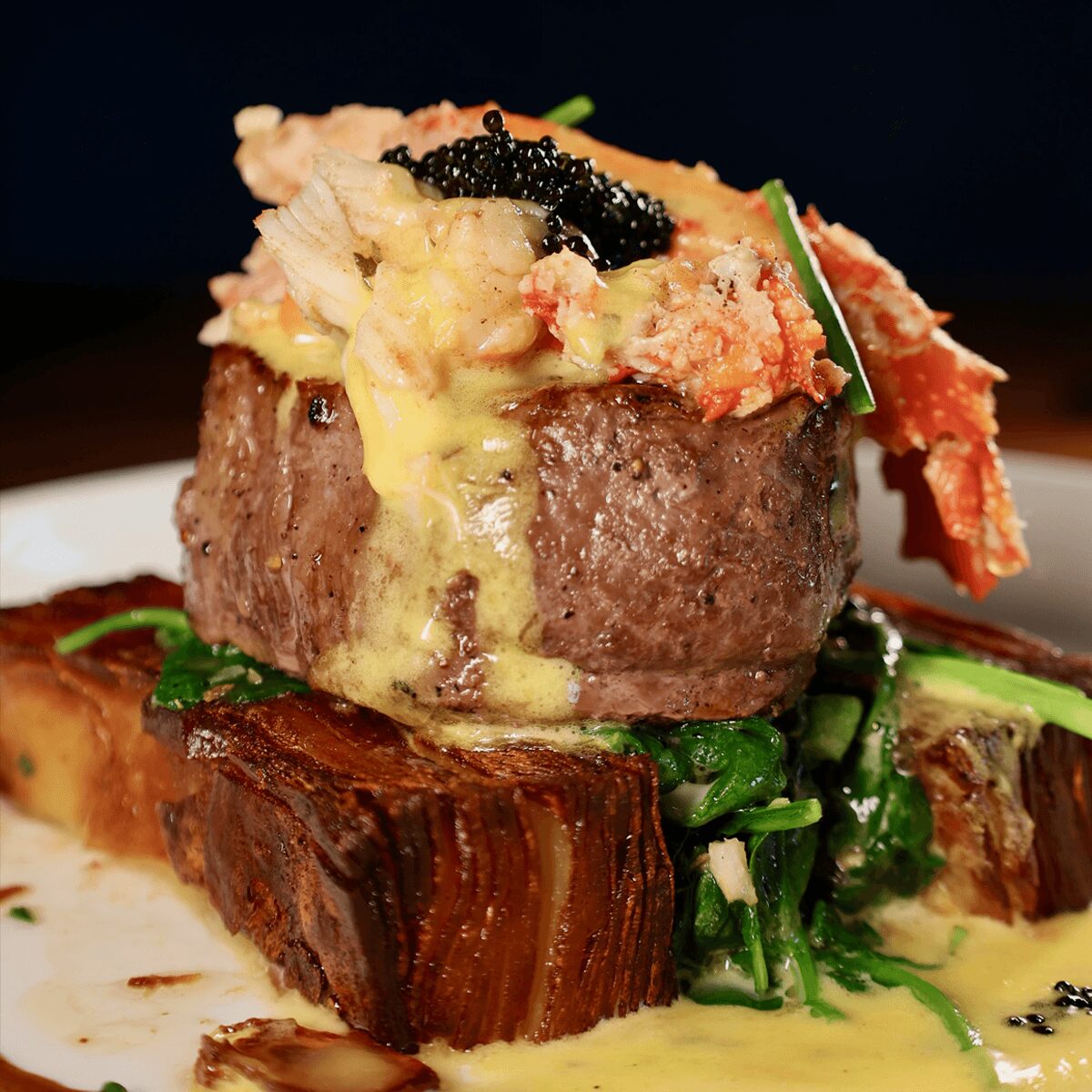 A gourmet dish with a beef patty topped with creamy sauce, caviar, and garnishes, served on a bed of greens and vegetables.