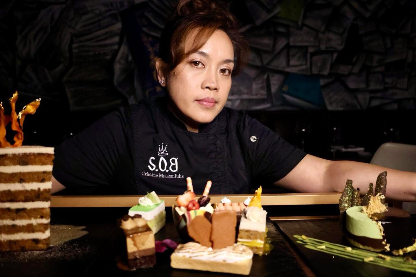 Woman with short hair wearing black shirt sitting at a table with assorted cakes and desserts.