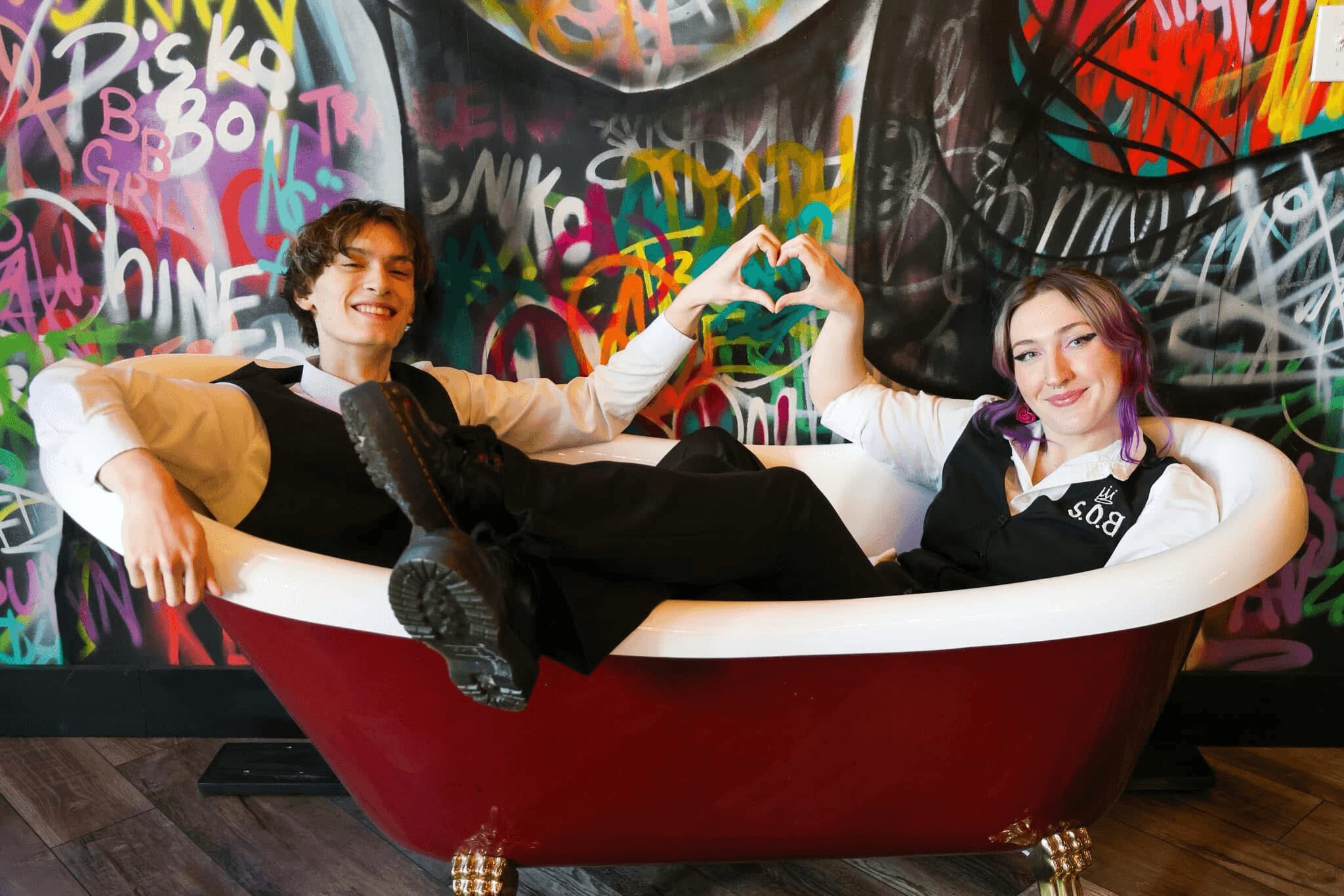 Two young people in a bathtub with graffiti-covered wall background, holding a green heart-shaped object, smiling.