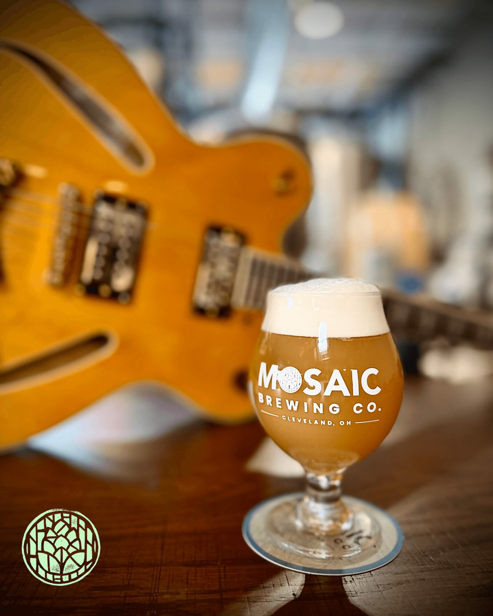 Glass of beer with foam on top, branded with Mosaic Brewing Co., on a wooden surface with a guitar in the background.