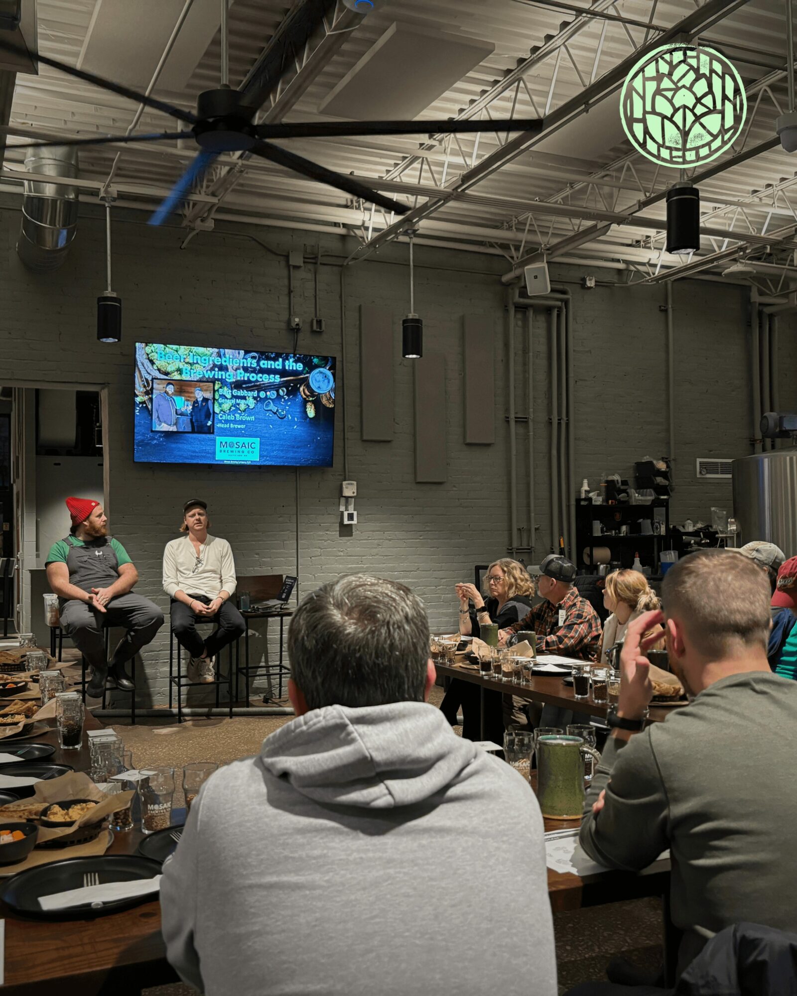 People seated at tables in a room with a large screen and a logo on the ceiling, listening to a presentation.