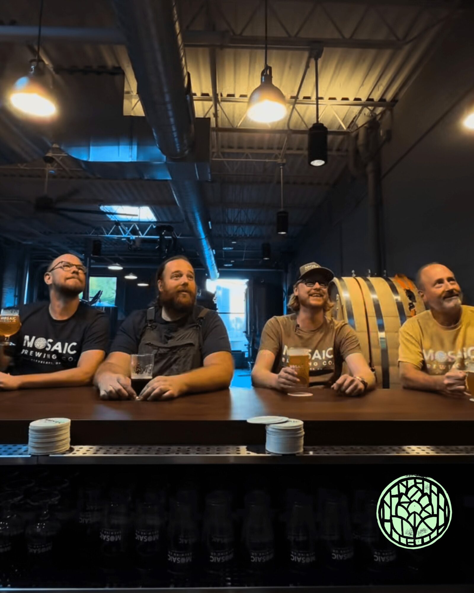 Four people sitting at a bar counter with beer glasses, barrels, and industrial ceiling lights above.