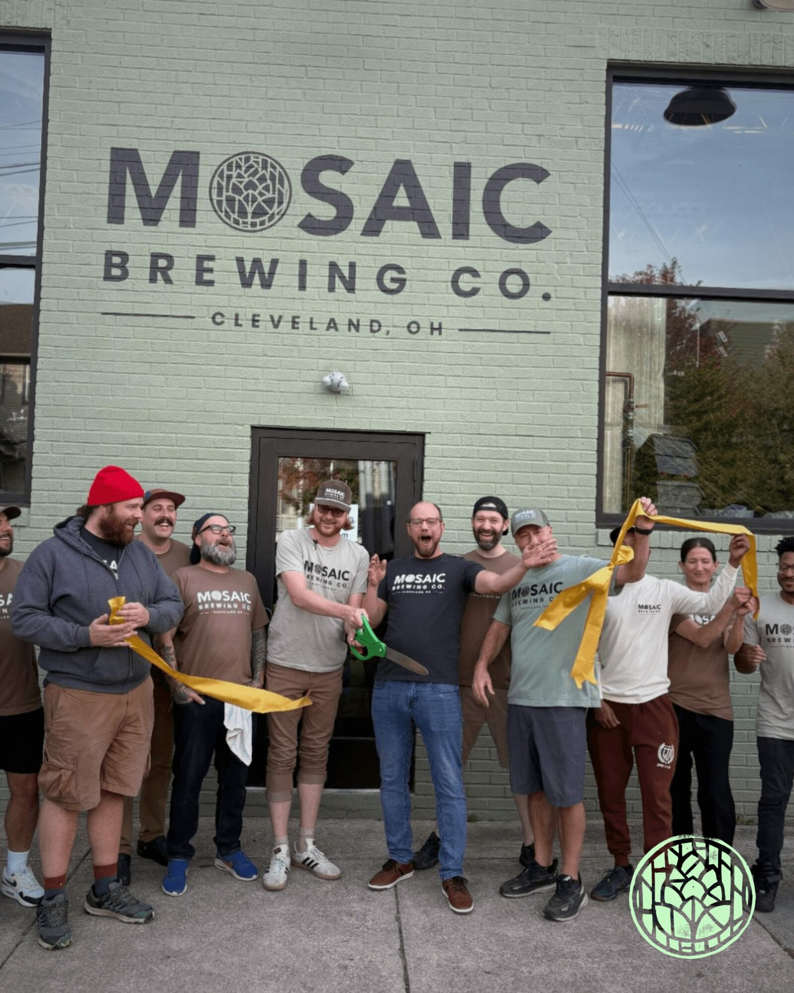 Group of people standing outside Mosaic Brewing Co. building, holding a yellow ribbon for a ribbon-cutting event.