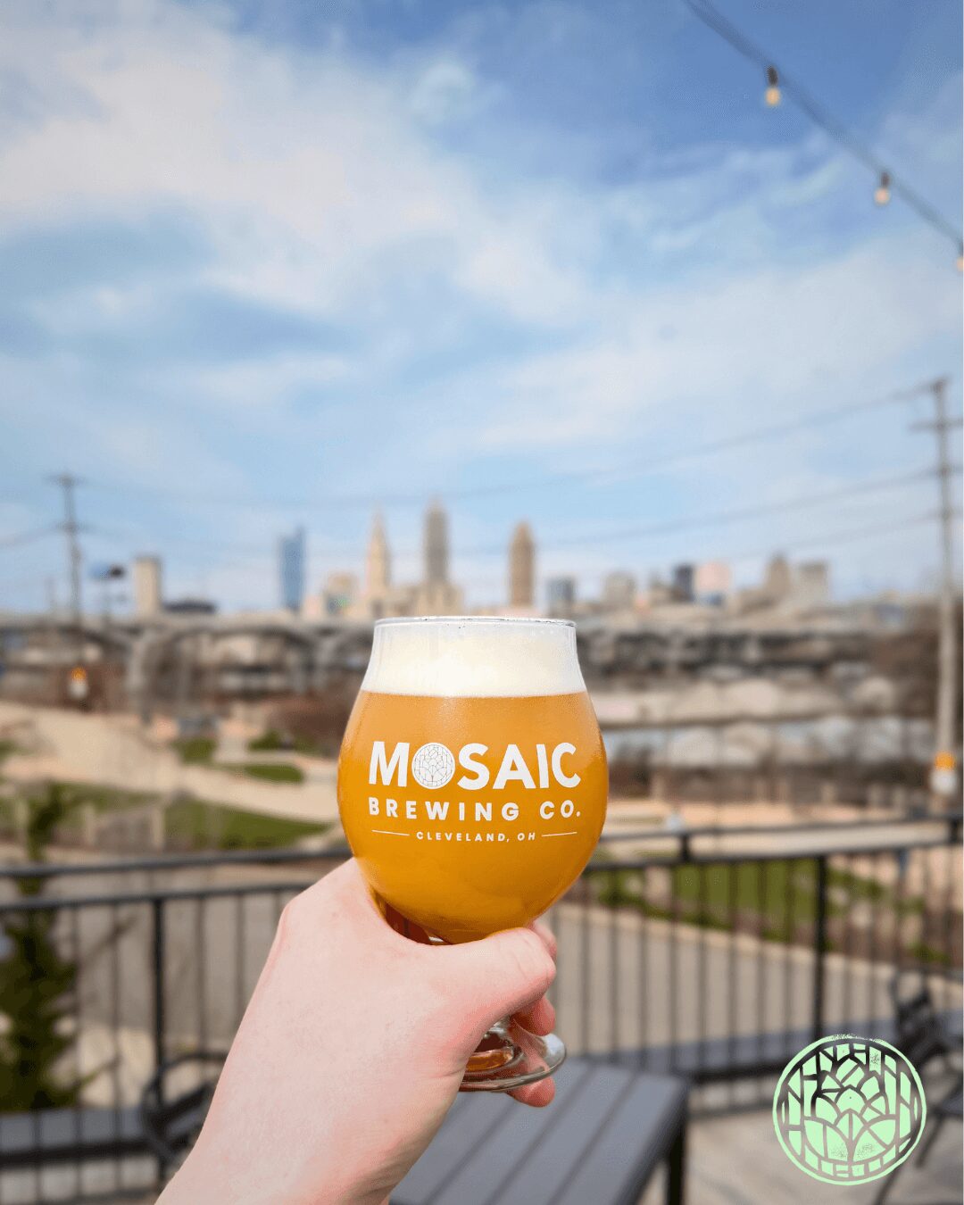 Hand holding a beer glass with orange and white logo, city skyline in background, blue sky with clouds.