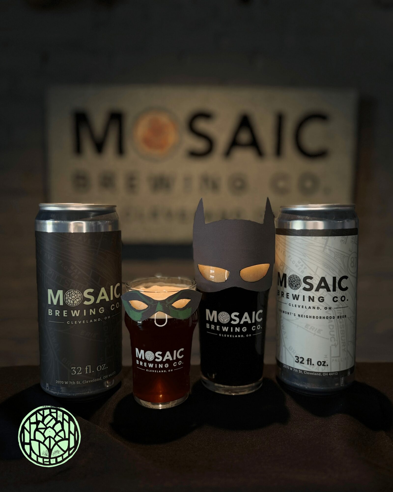 Two cans, a glass, and a black container with a logo in front of a blurred sign reading MOSAIC BREWING CO.