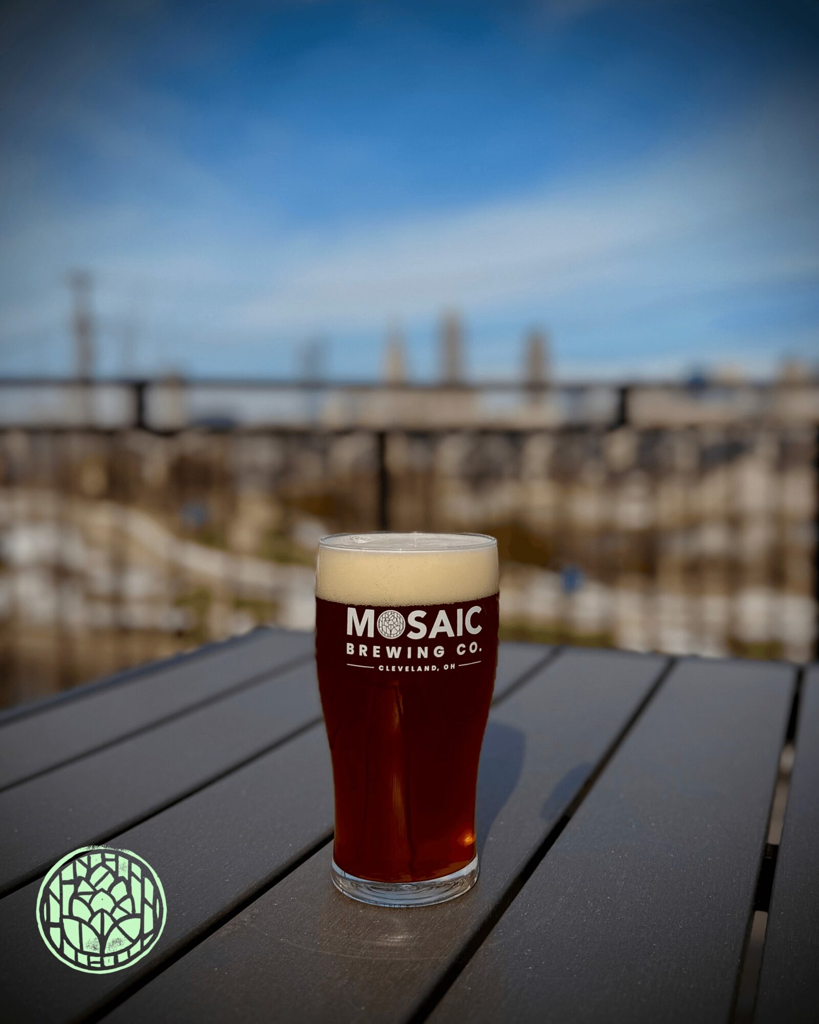 A glass of dark beer with foam on top on a black outdoor table, cityscape background, blue sky.