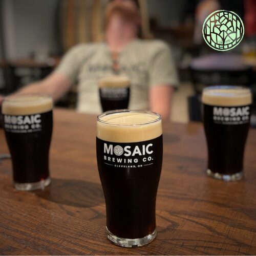Three glasses of dark beer on a wooden table with a person in the background.