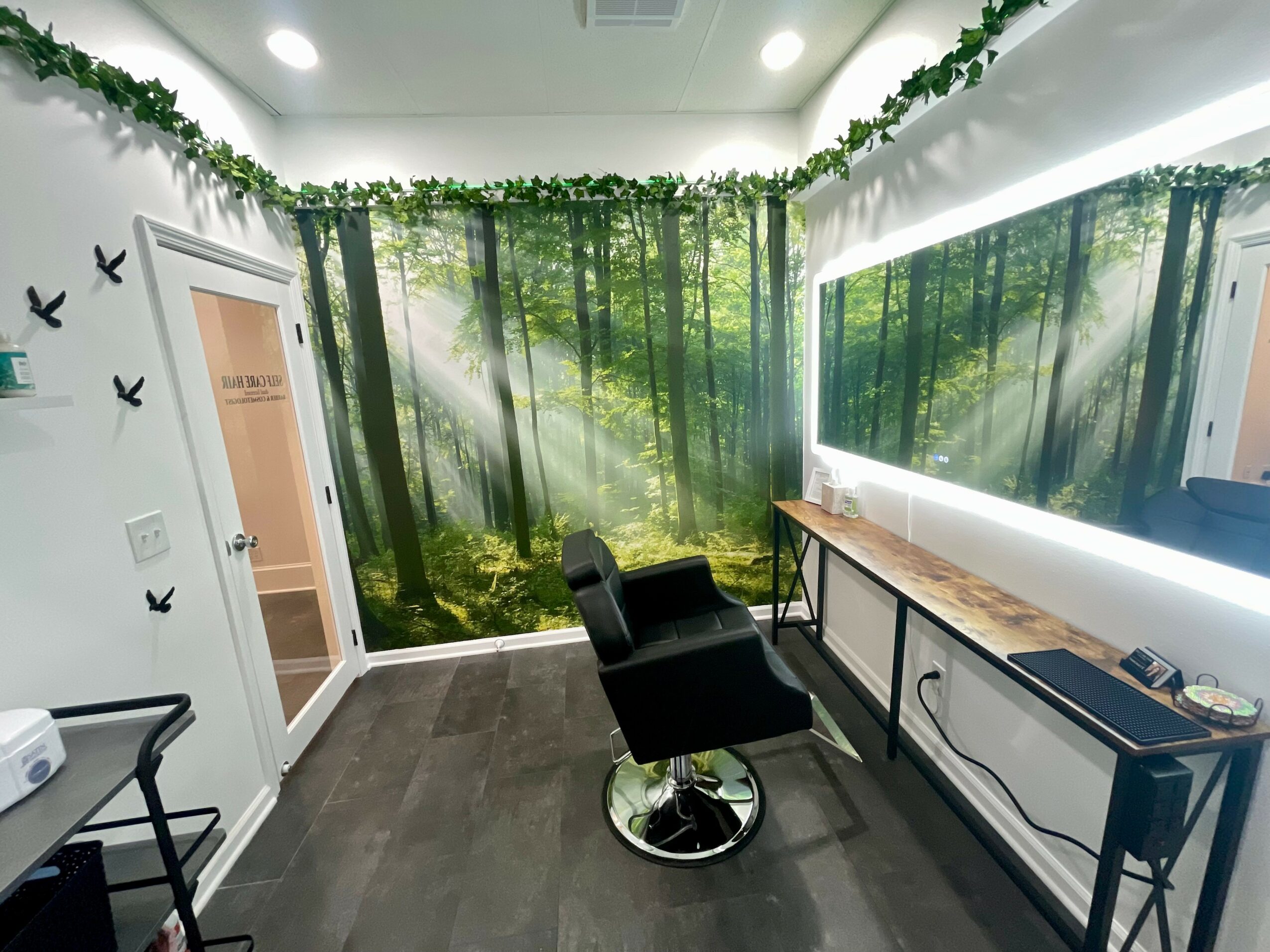 Room with forest-themed wall mural, black chair, wooden desk, and window with white blinds, decorated with green foliage garland.