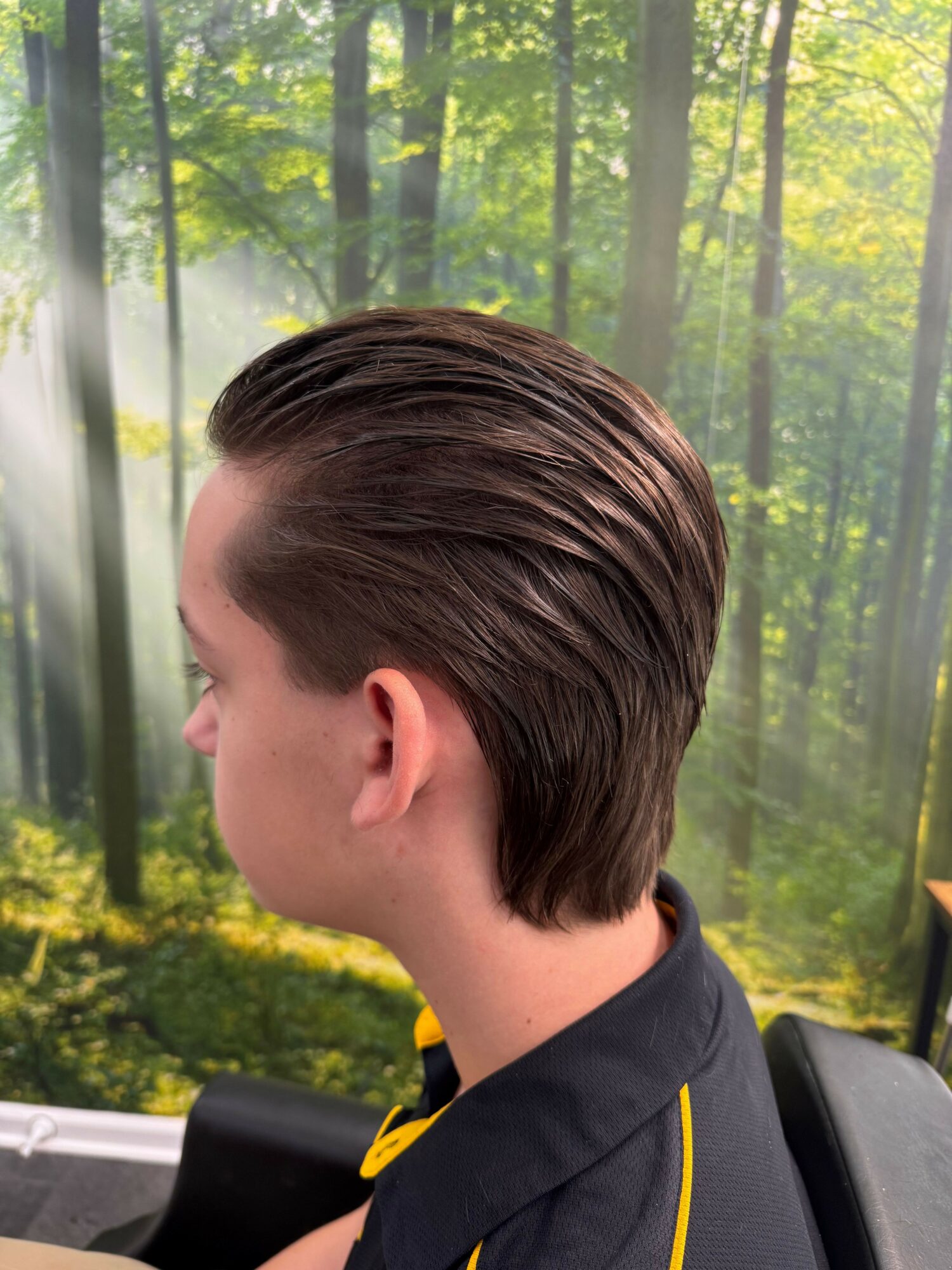 Side view of a young person with slicked-back hair, wearing a dark shirt, in front of a forest background.