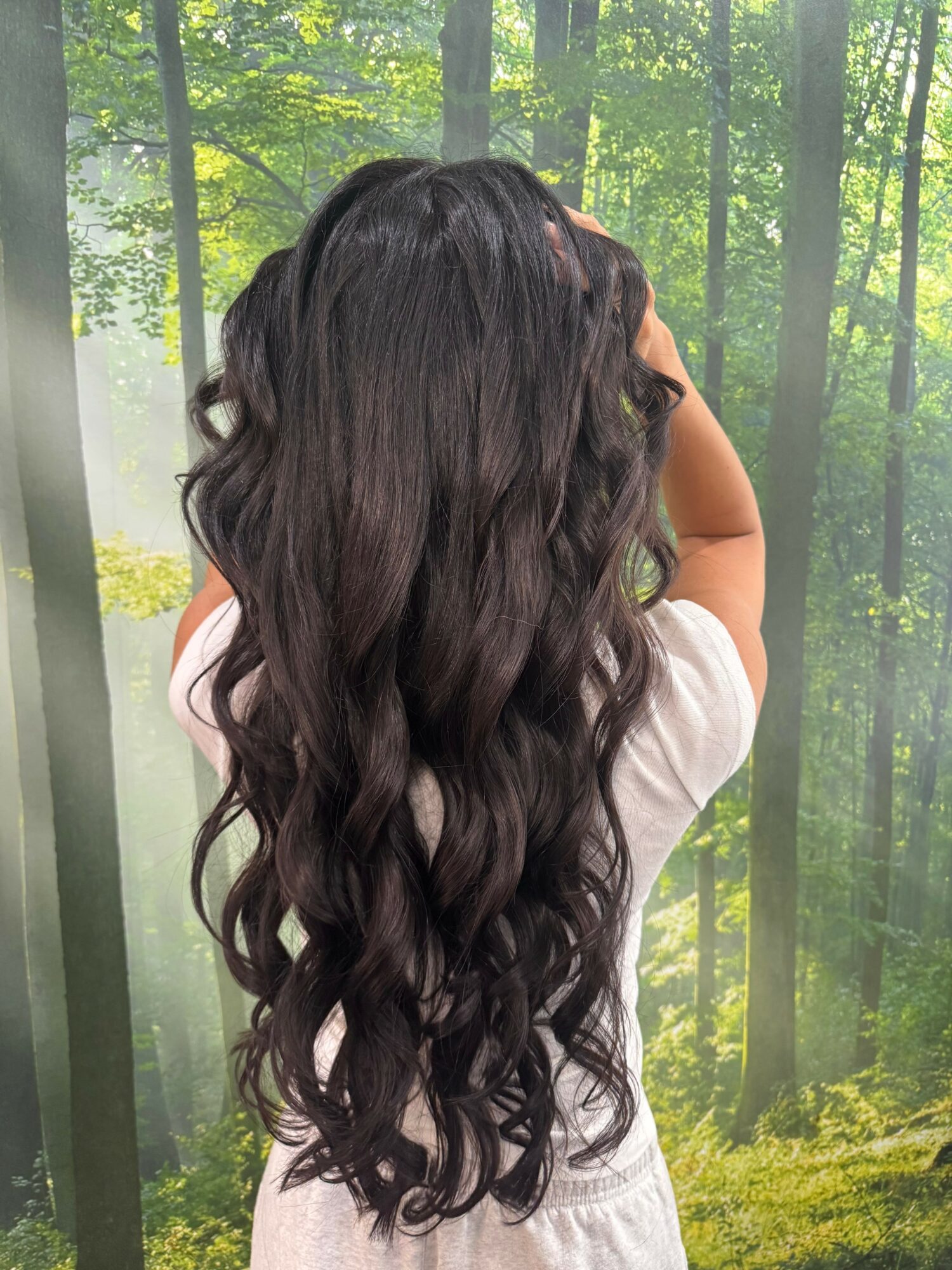 Person with long, wavy dark hair standing outdoors with trees in the background, facing away from the camera.