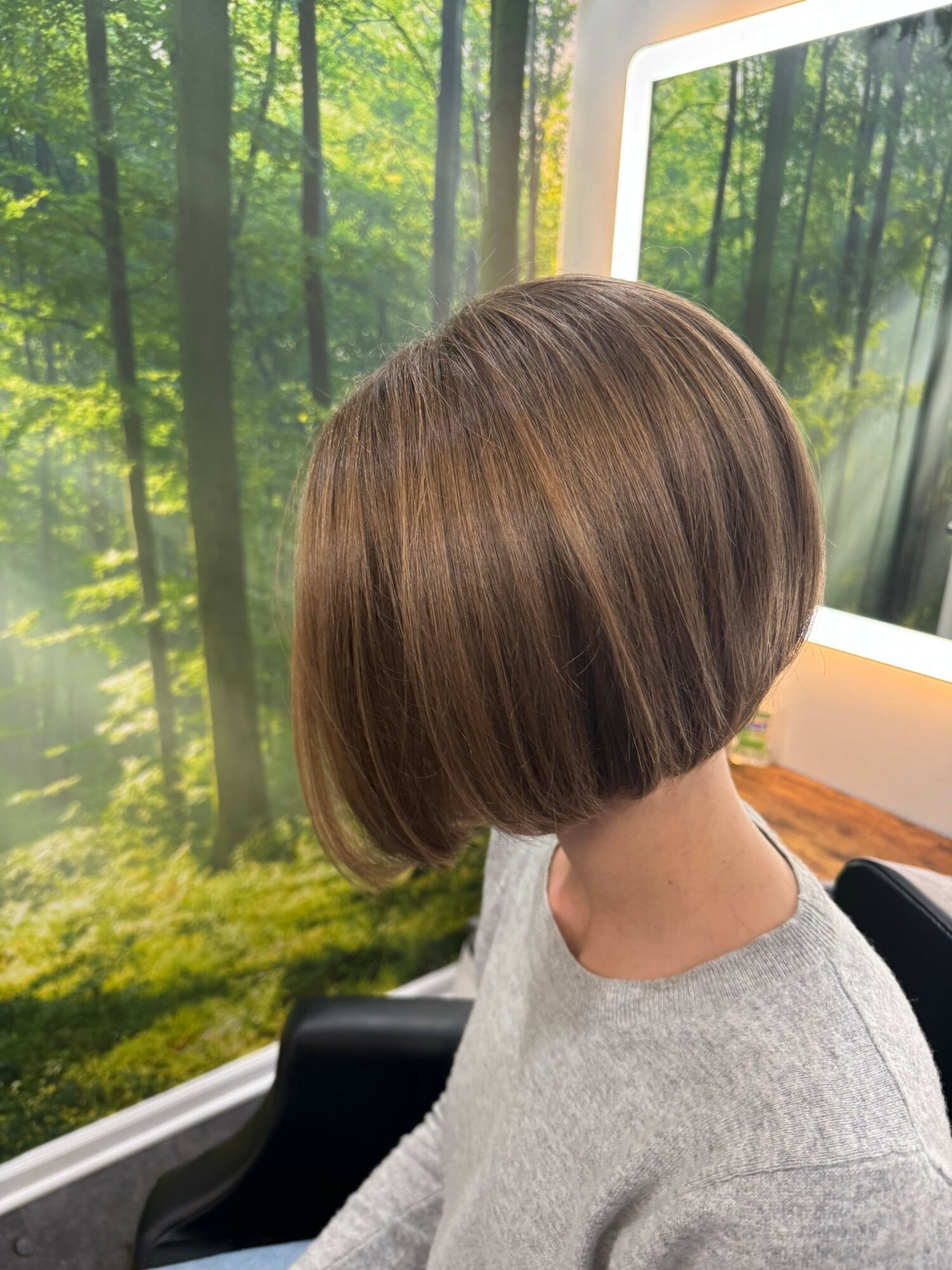 Person with short, straight, brown hair sitting in a chair near a window with a forest view.