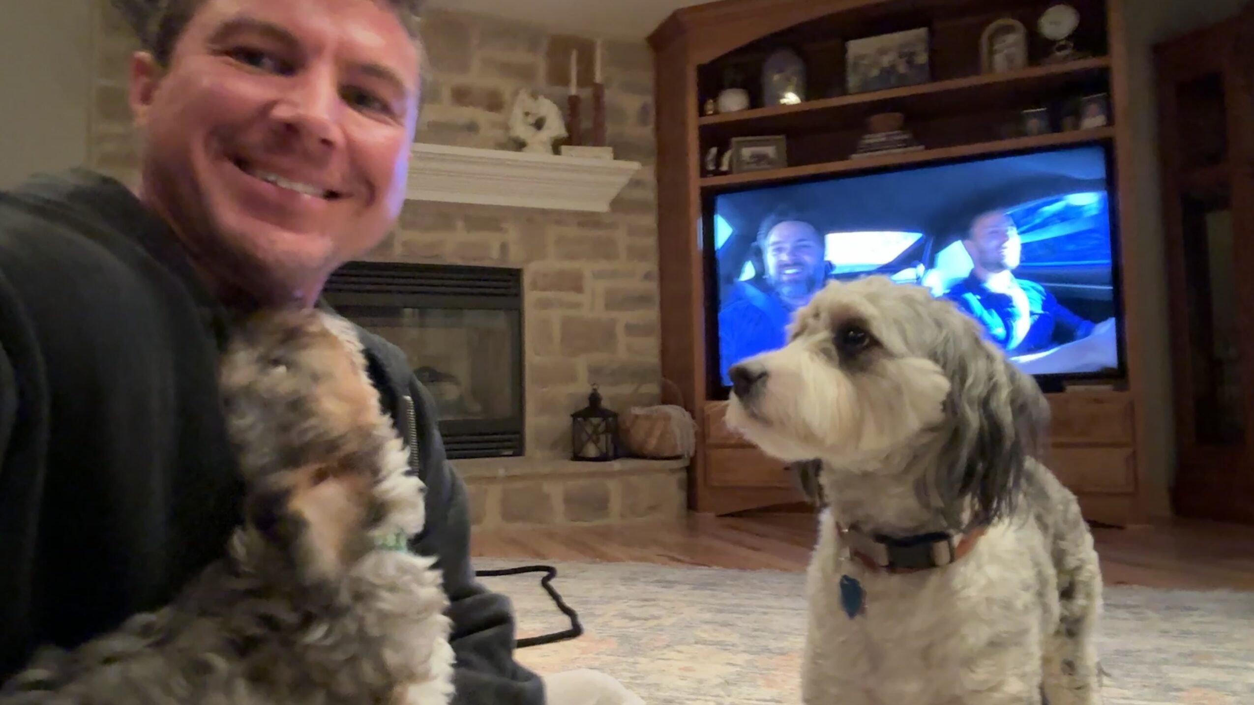 Person smiling holding a small dog, with a larger dog sitting nearby in a living room. TV in background.