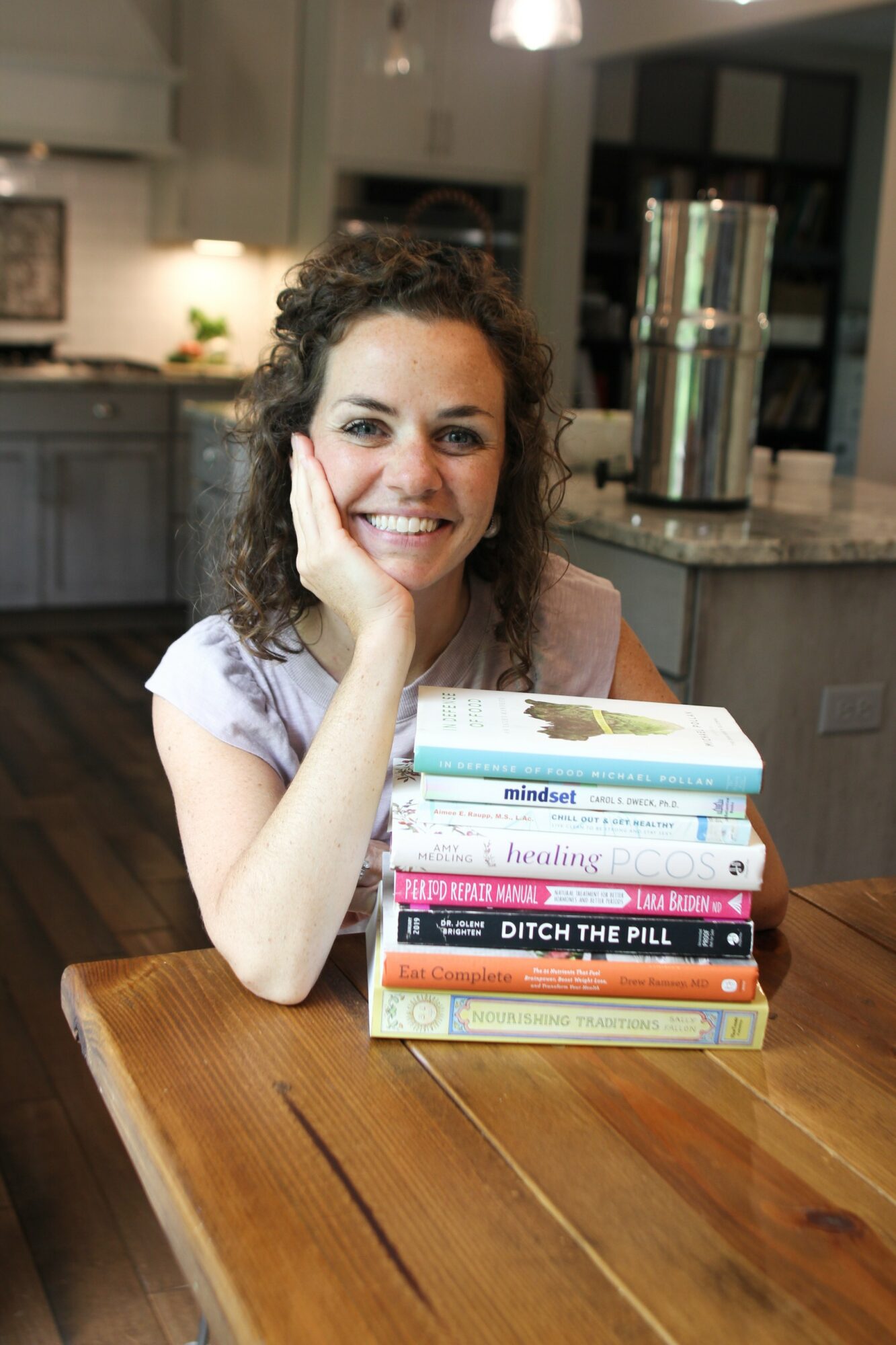Woman with curly hair smiling, resting her chin on her hand, sitting at a wooden table with a stack of books.