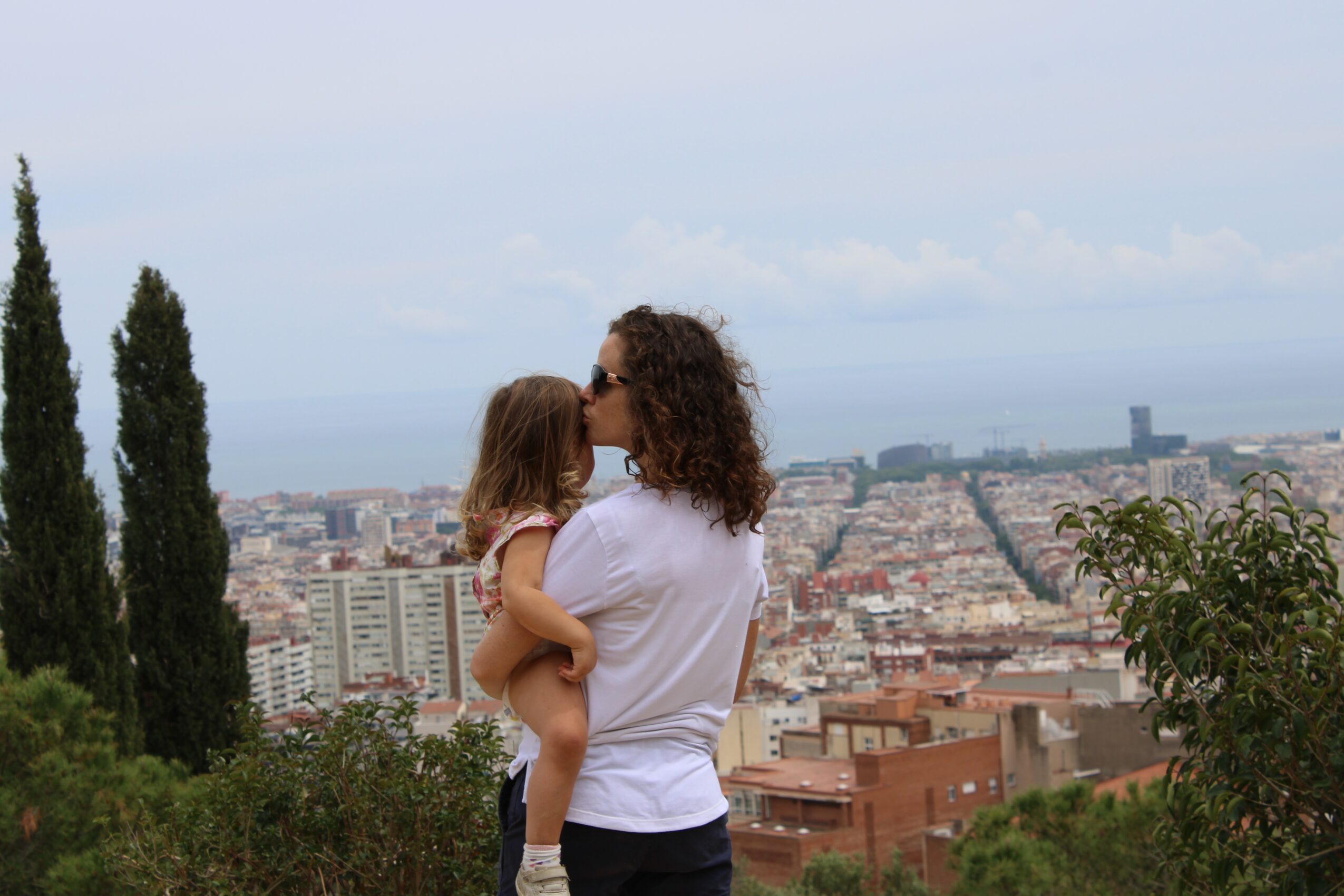 Woman holding a child, both kissing, with cityscape and trees in the background.