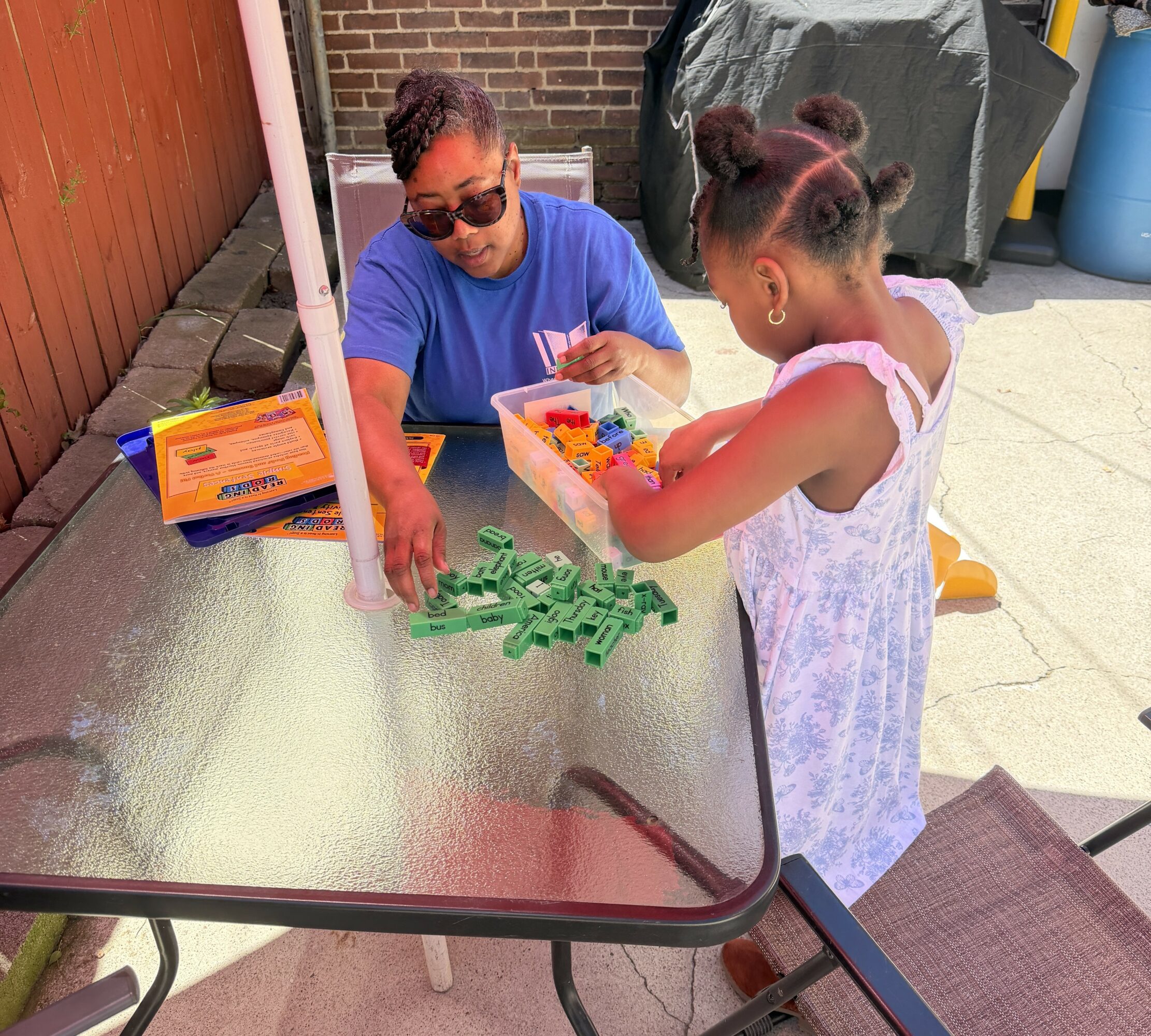 Two children playing with colorful plastic building blocks at an outdoor table, one girl in a white dress, one boy in a blue shirt, on a patio.