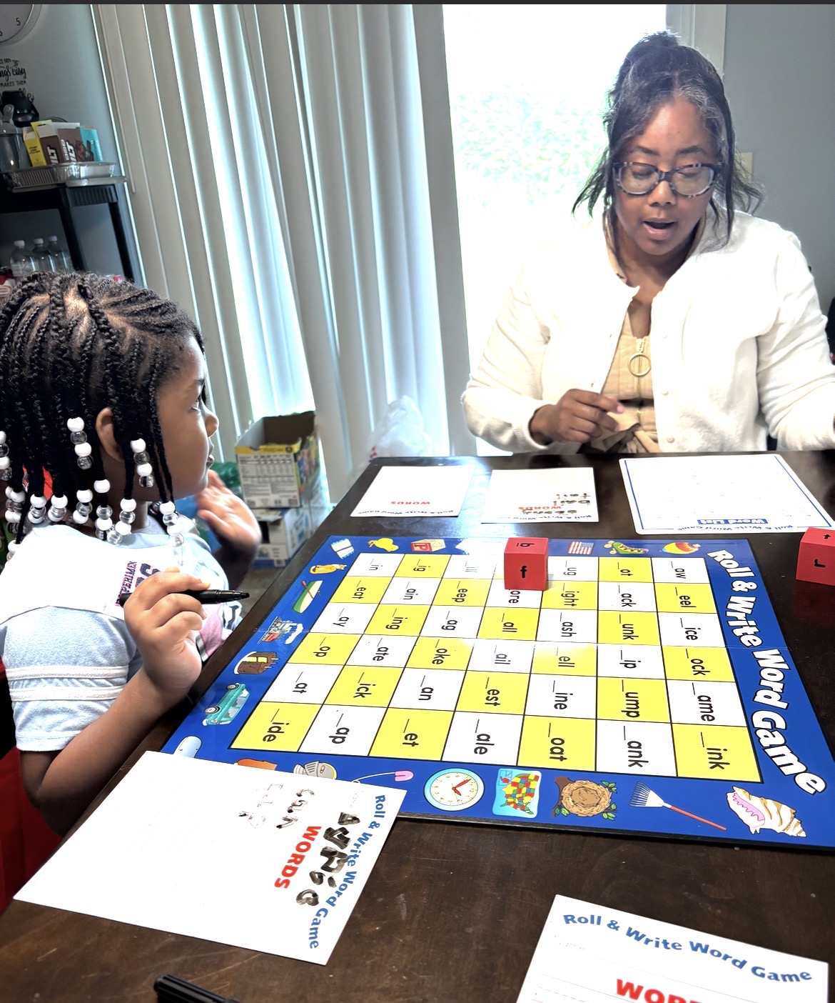 A woman and a young girl play a word game at a table indoors, with a game board, cards, and a die visible.