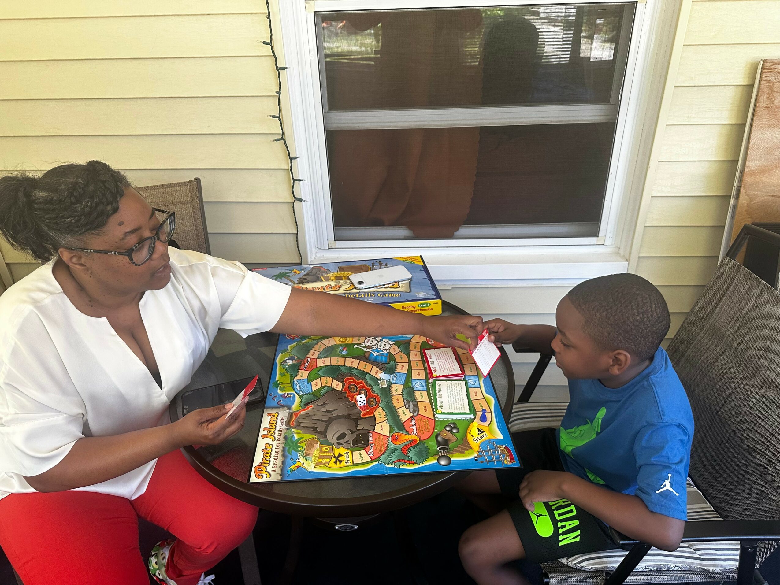 Woman and boy playing a board game on a table outside a house, with a window behind them.