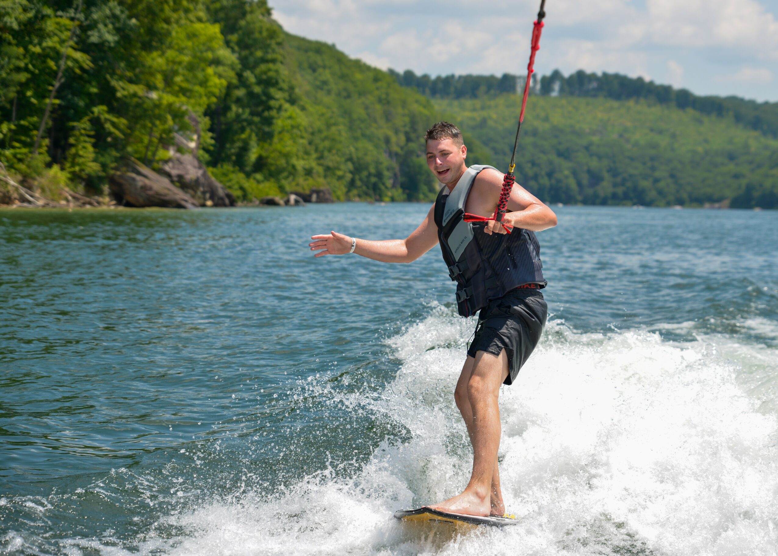 Person wakeboarding on water, holding a handle, with trees and hills in the background.