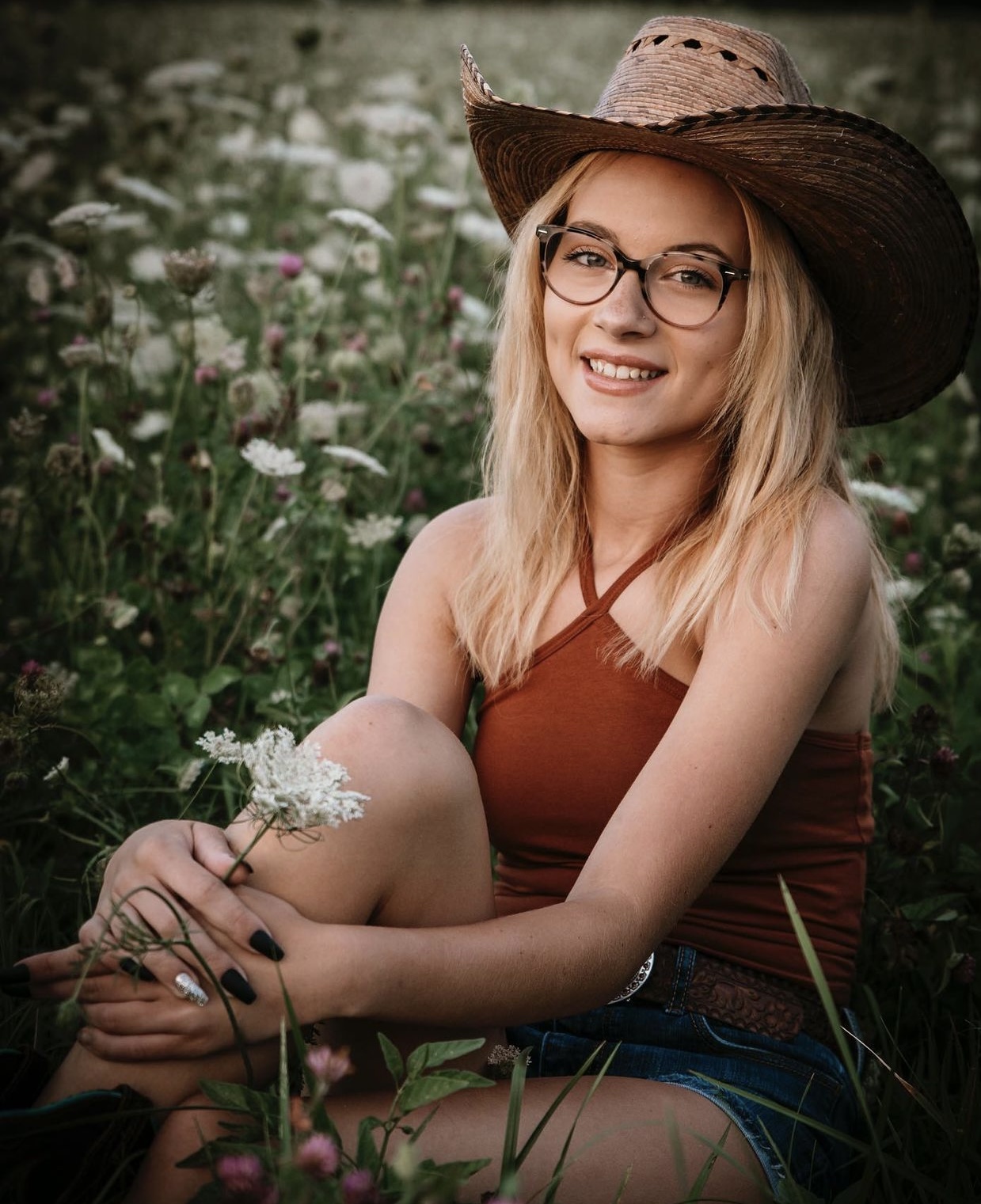 Young woman with glasses and a wide-brimmed hat sitting in a field of flowers, smiling, wearing a sleeveless top and shorts.