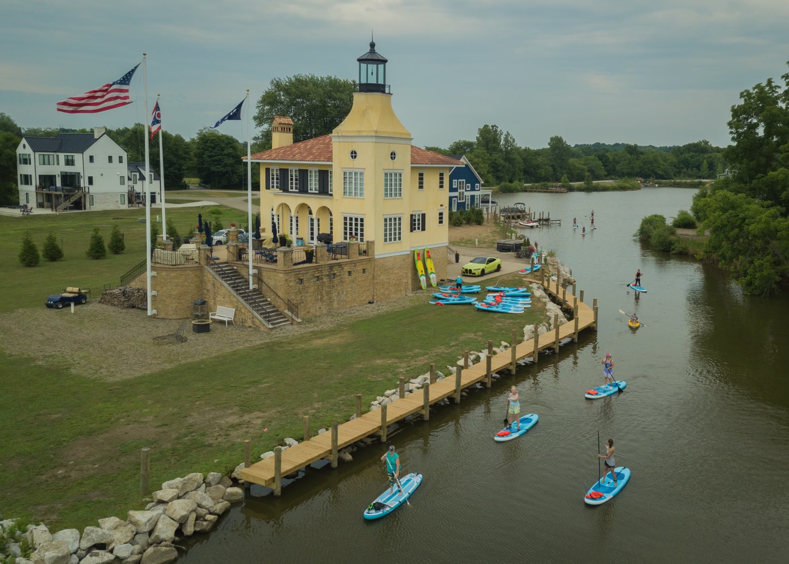 Lighthouse-style building with a dock, boats on water, and flags, near a river with trees and buildings in background.