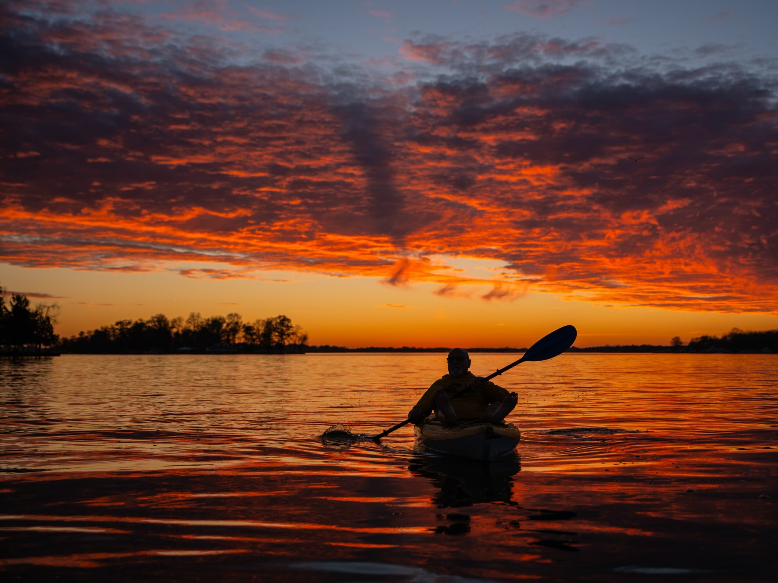 Person kayaking on water during sunset with colorful sky and clouds, silhouette visible, calm water reflections.