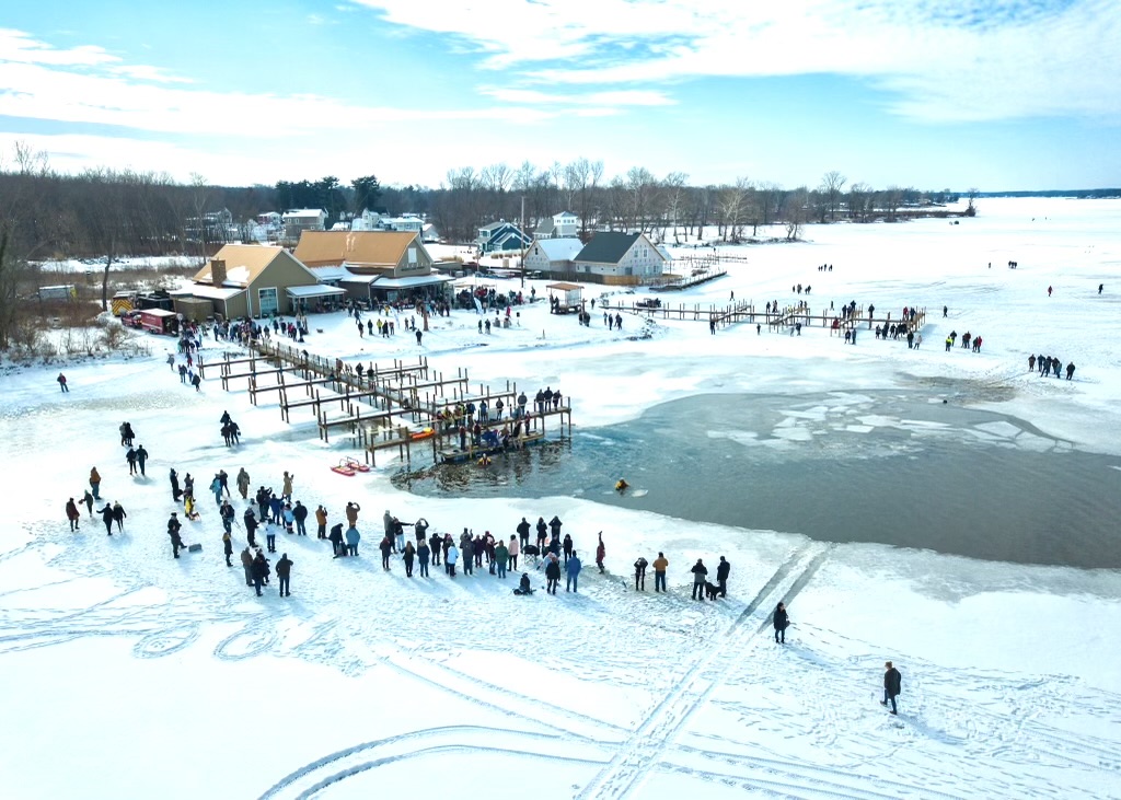People skating and walking on a snowy frozen lake with buildings and trees in the background.