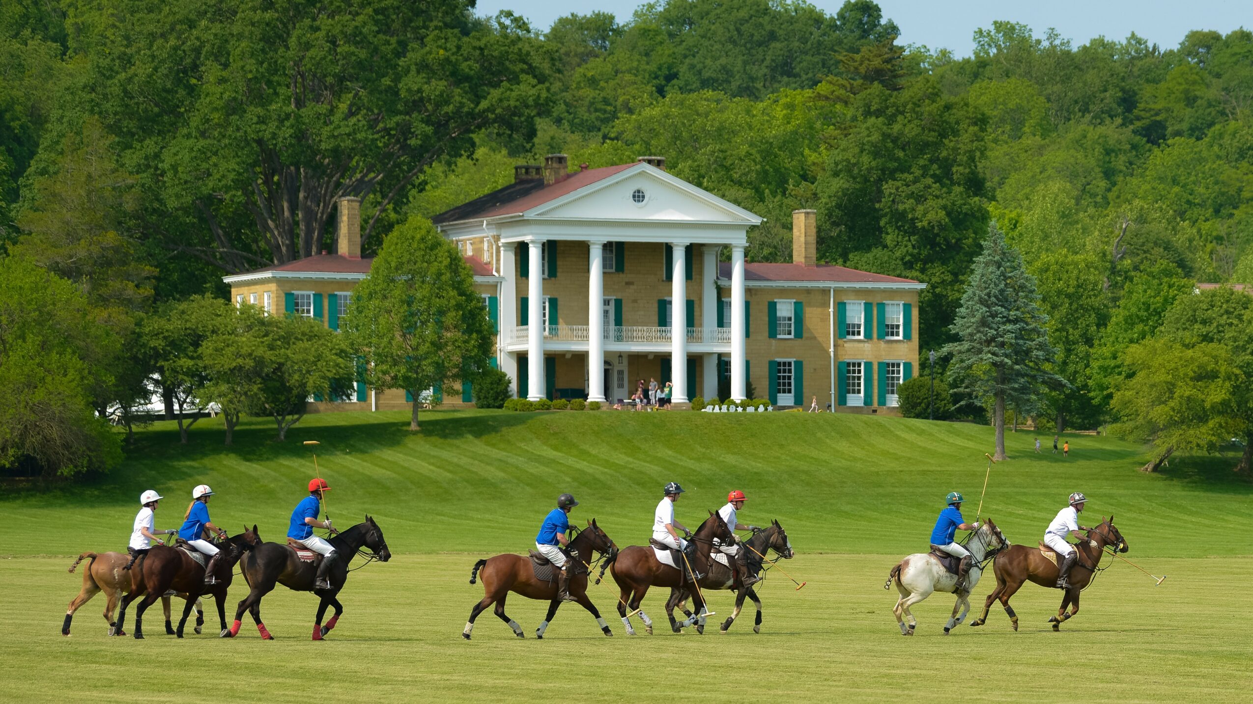 Group of people riding horses on a grassy field with a large house and trees in the background.
