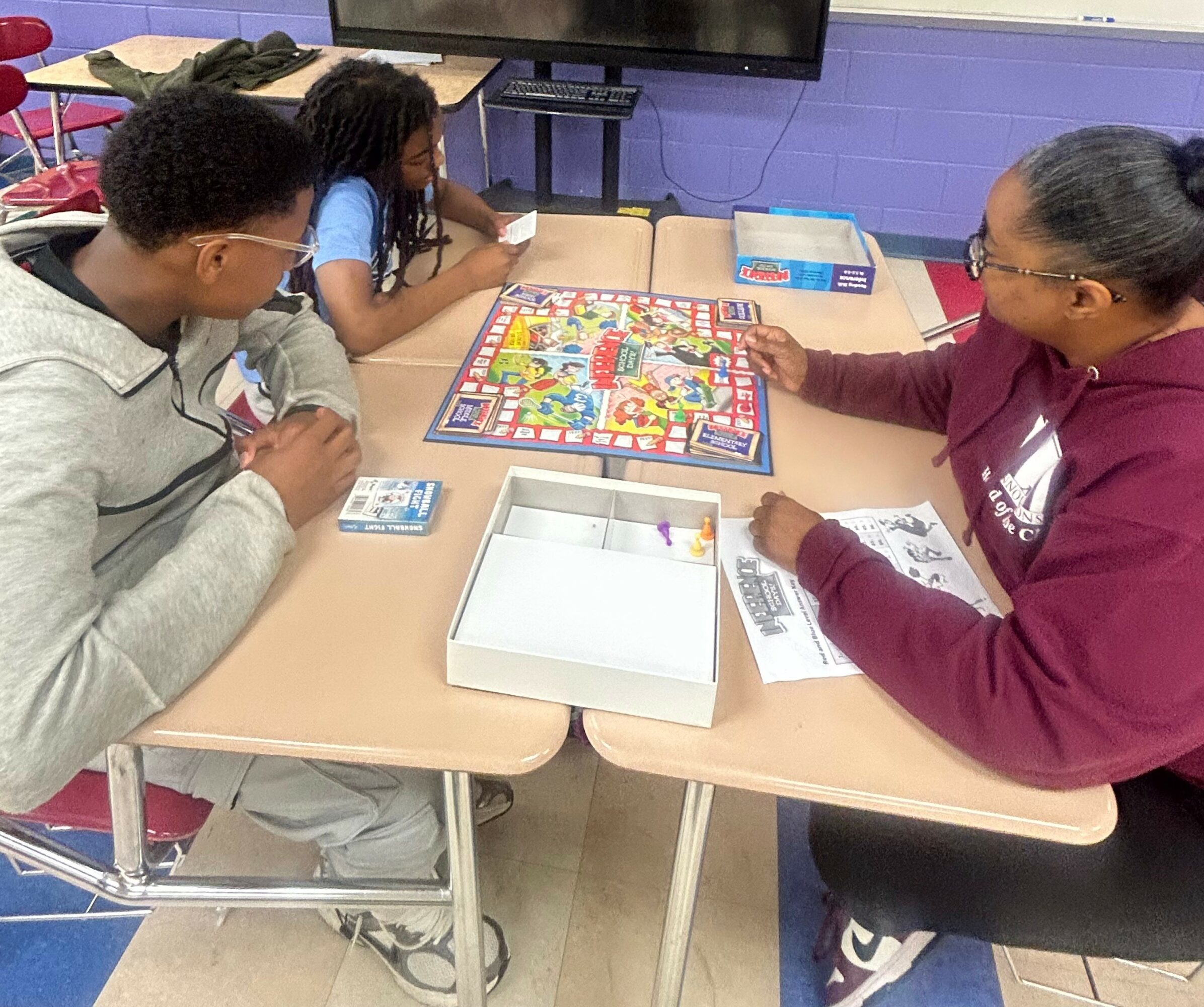 Three people playing a colorful board game at a table in a classroom, with game pieces and cards visible.