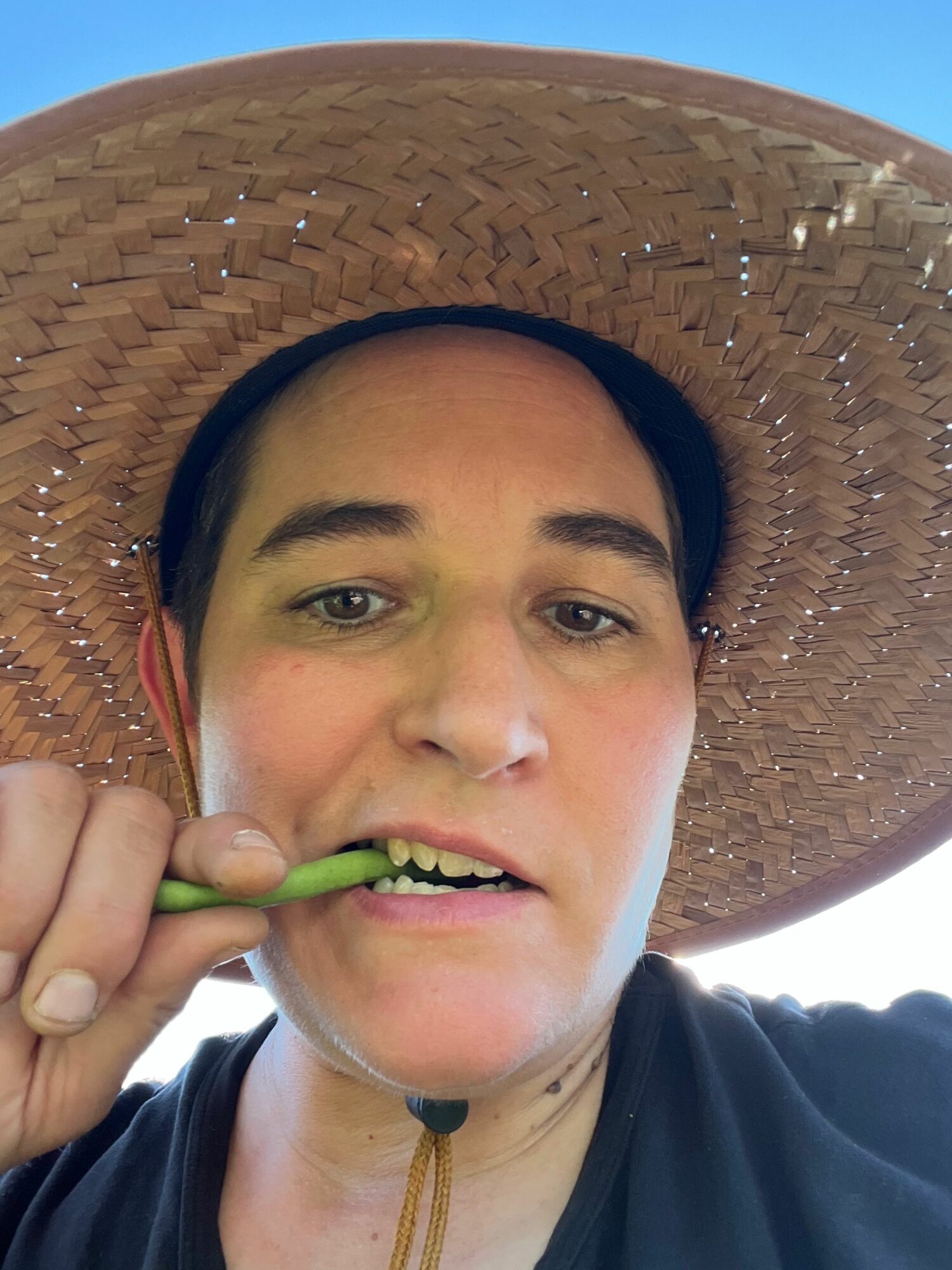 Person wearing a large straw hat, brushing teeth outdoors, holding a green toothbrush, with a clear sky background.