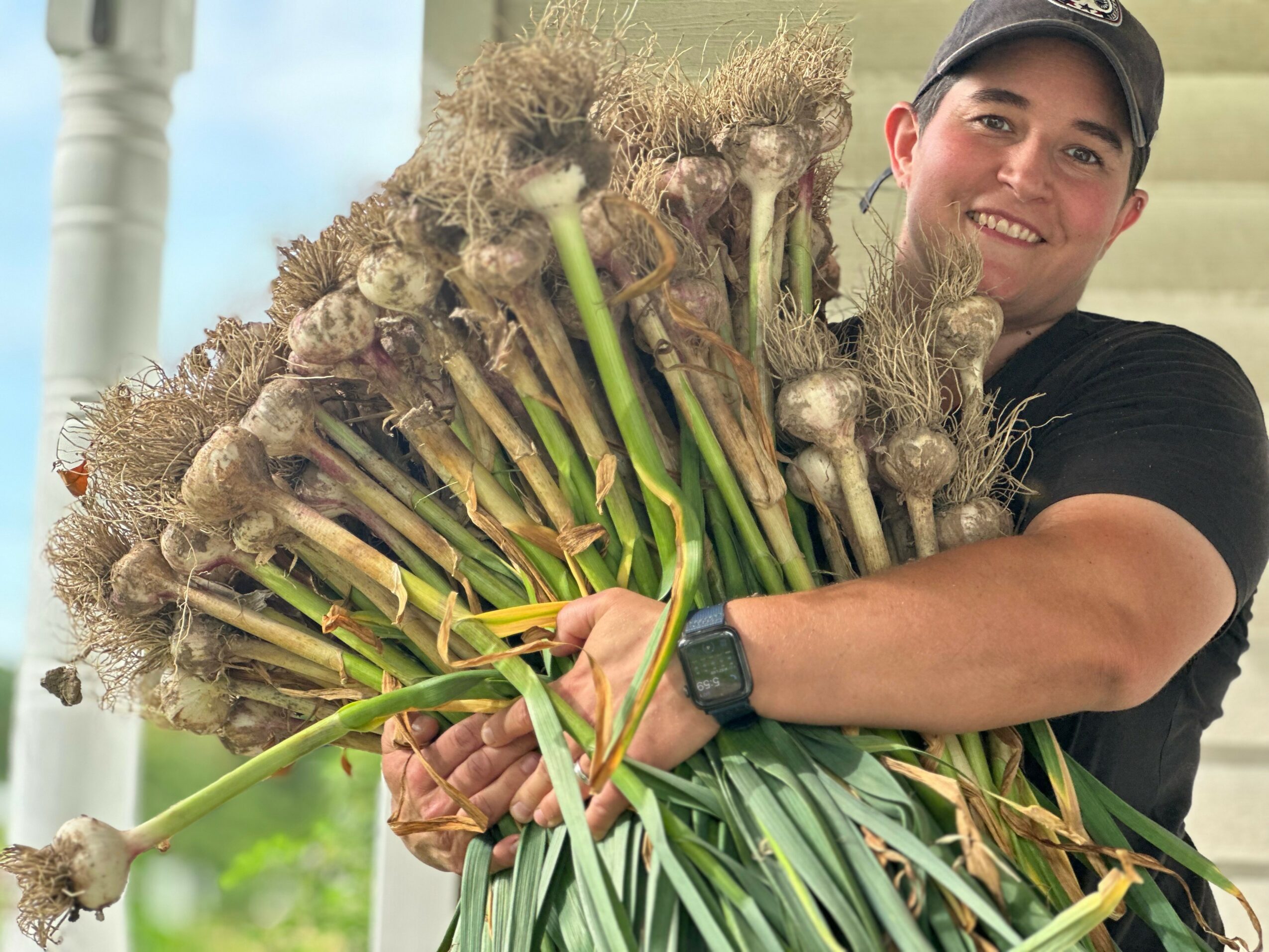 Person holding a large bunch of freshly harvested asparagus outdoors, smiling, wearing a black shirt and cap.