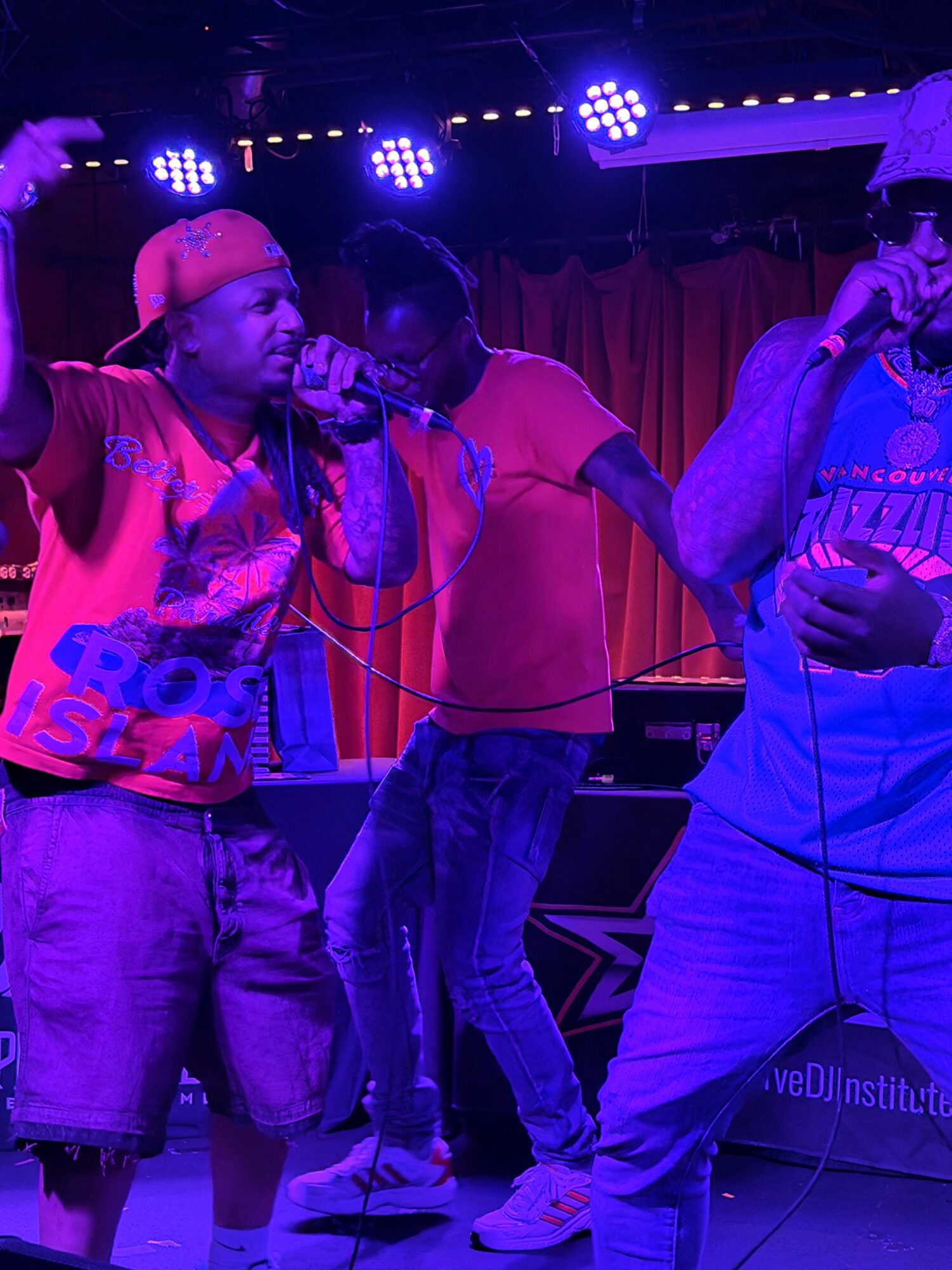 Two people on stage, one holding a microphone, with colorful lighting and curtains in the background.