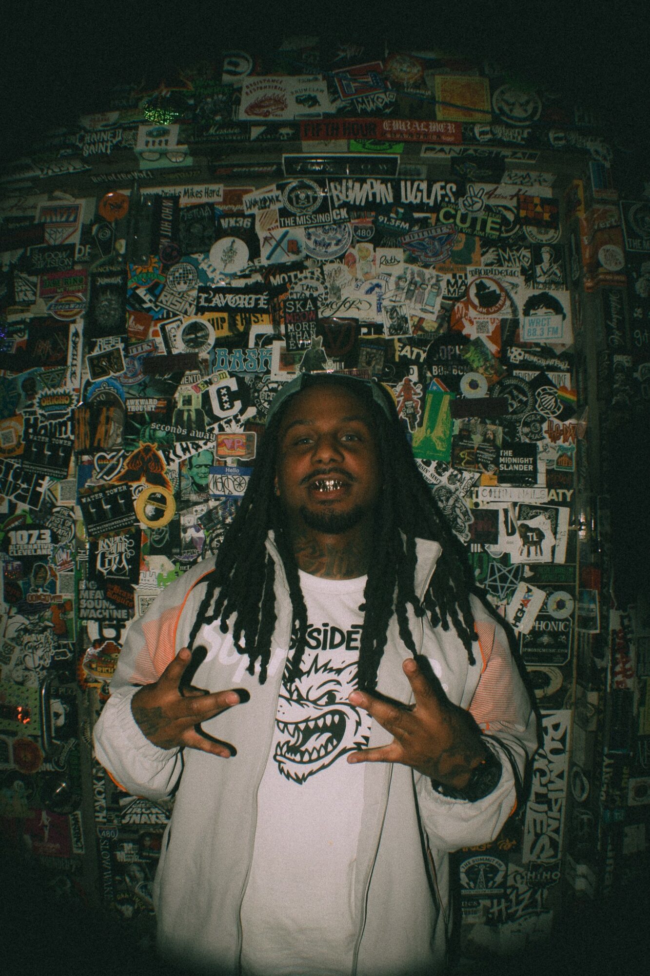 Person with long dreadlocks wearing a white jacket and graphic t-shirt, standing in front of a wall covered with stickers and posters.