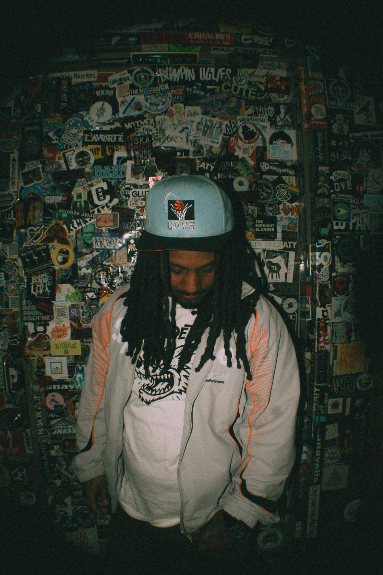 Person with dreadlocks wearing a white jacket and helmet, standing in front of a wall covered with stickers and posters.
