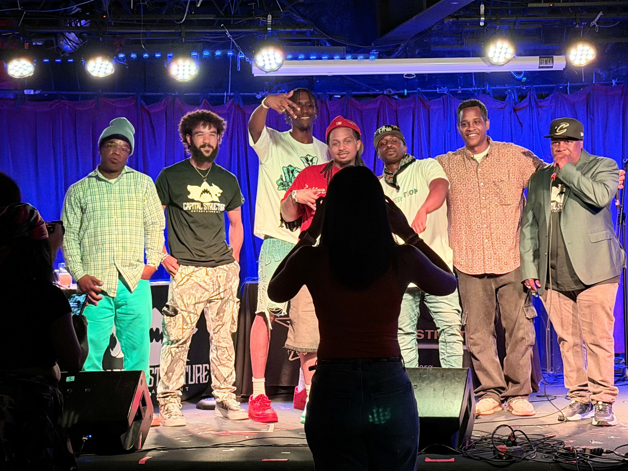 Group of seven people on stage with a woman in the foreground taking a photo, stage lights above, blue curtain background.