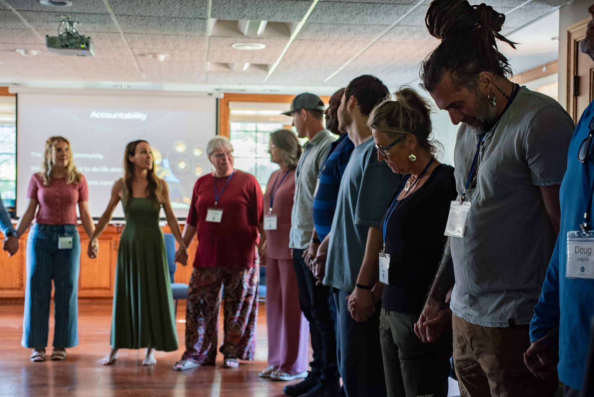 Group of people standing in a circle indoors, some with name tags, participating in a group activity or discussion.
