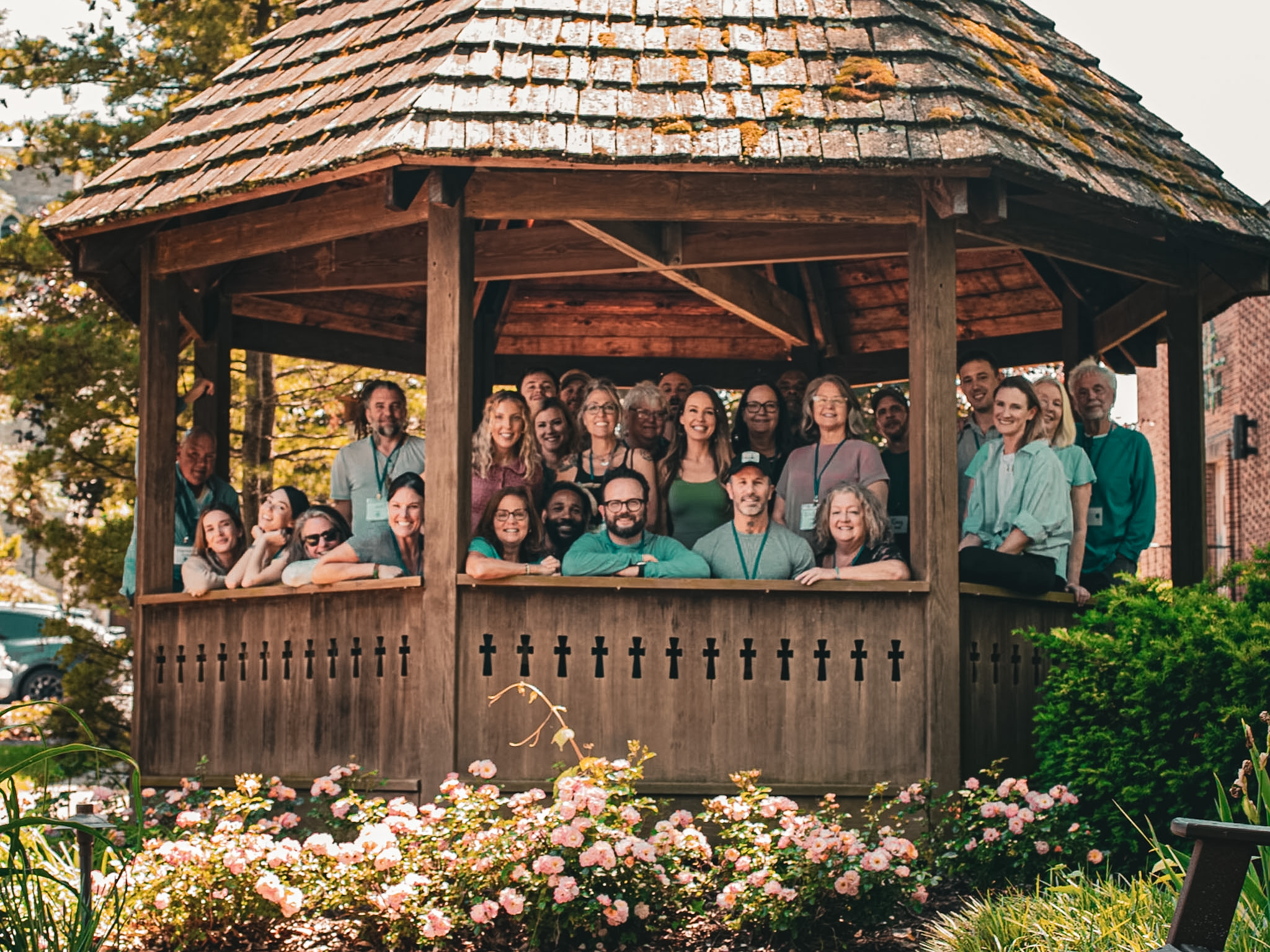 Group of people gathered on a wooden gazebo with a shingled roof, surrounded by flowers and greenery.