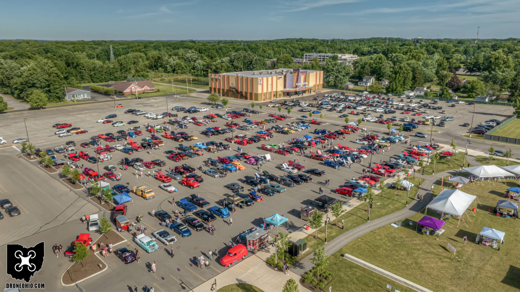 A large parking lot filled with cars, surrounded by trees and a building in the background, with tents set up nearby.