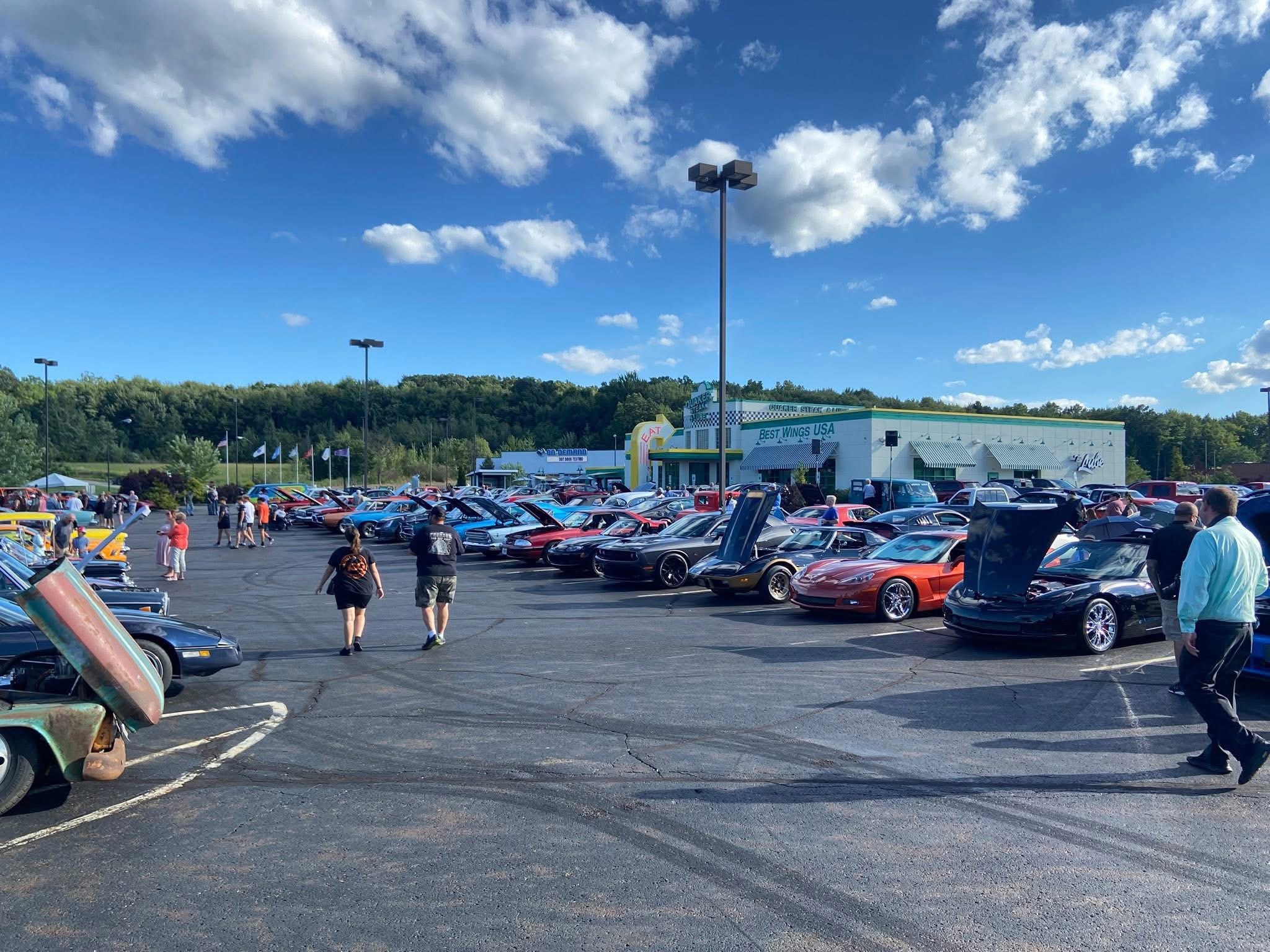Car show in parking lot with many vehicles, some with open hoods, under a blue sky with clouds.