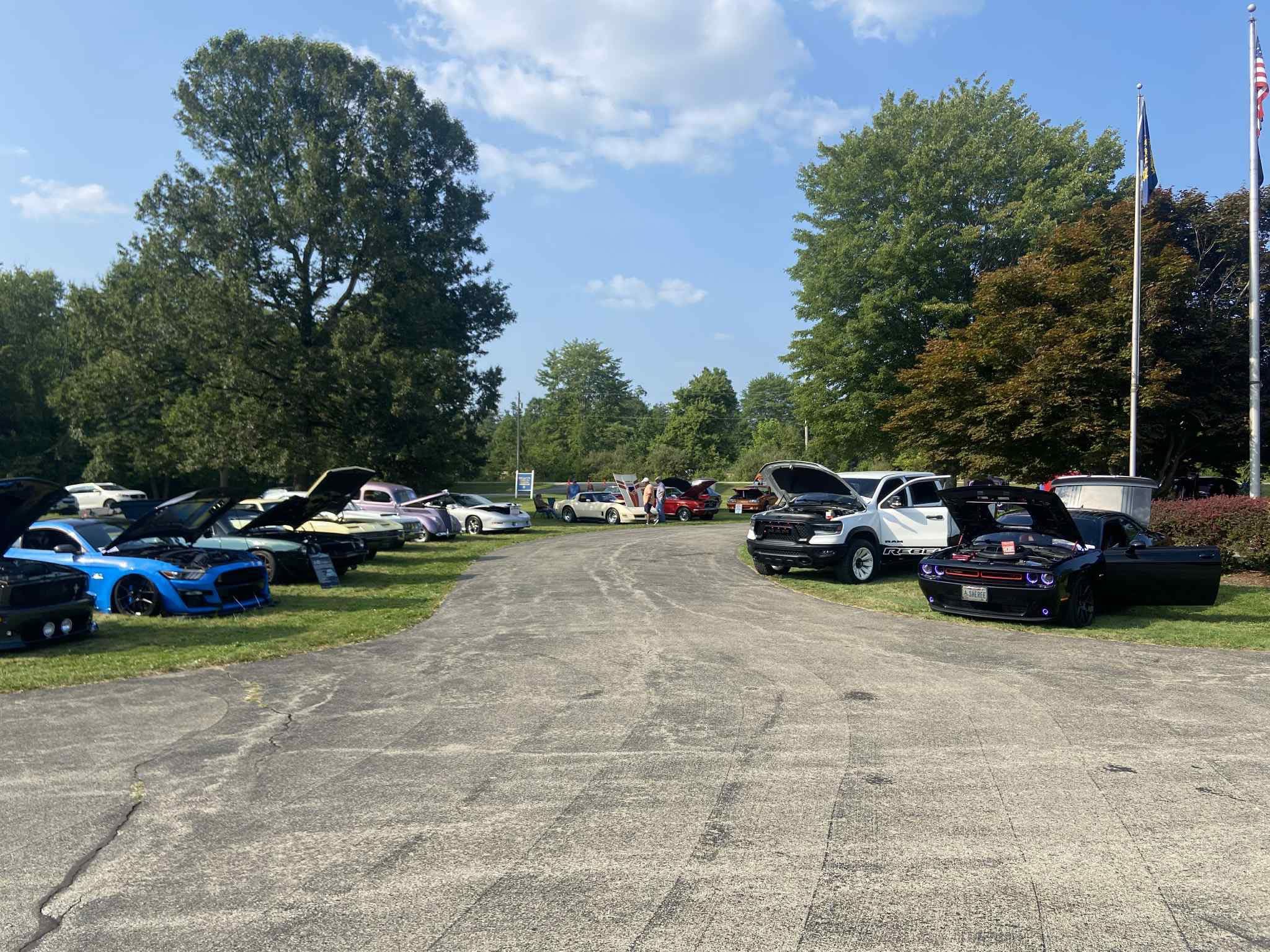Parking lot with cars on both sides, trees in background, blue sky with clouds, and flags on poles.