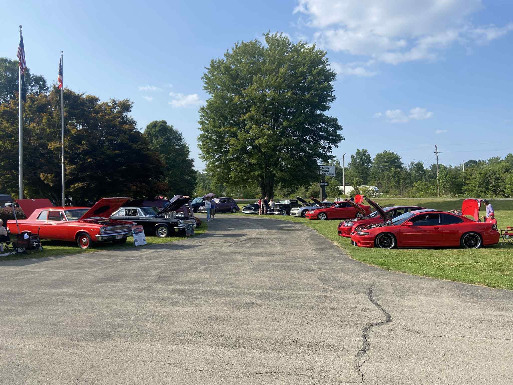 Car show with various vehicles parked on grass and pavement, large tree in background, flags on poles, blue sky with clouds.