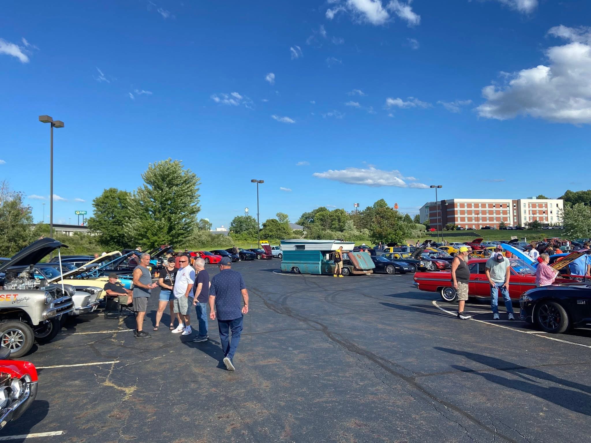 People gather around cars in a parking lot on a sunny day with a blue sky and scattered clouds.