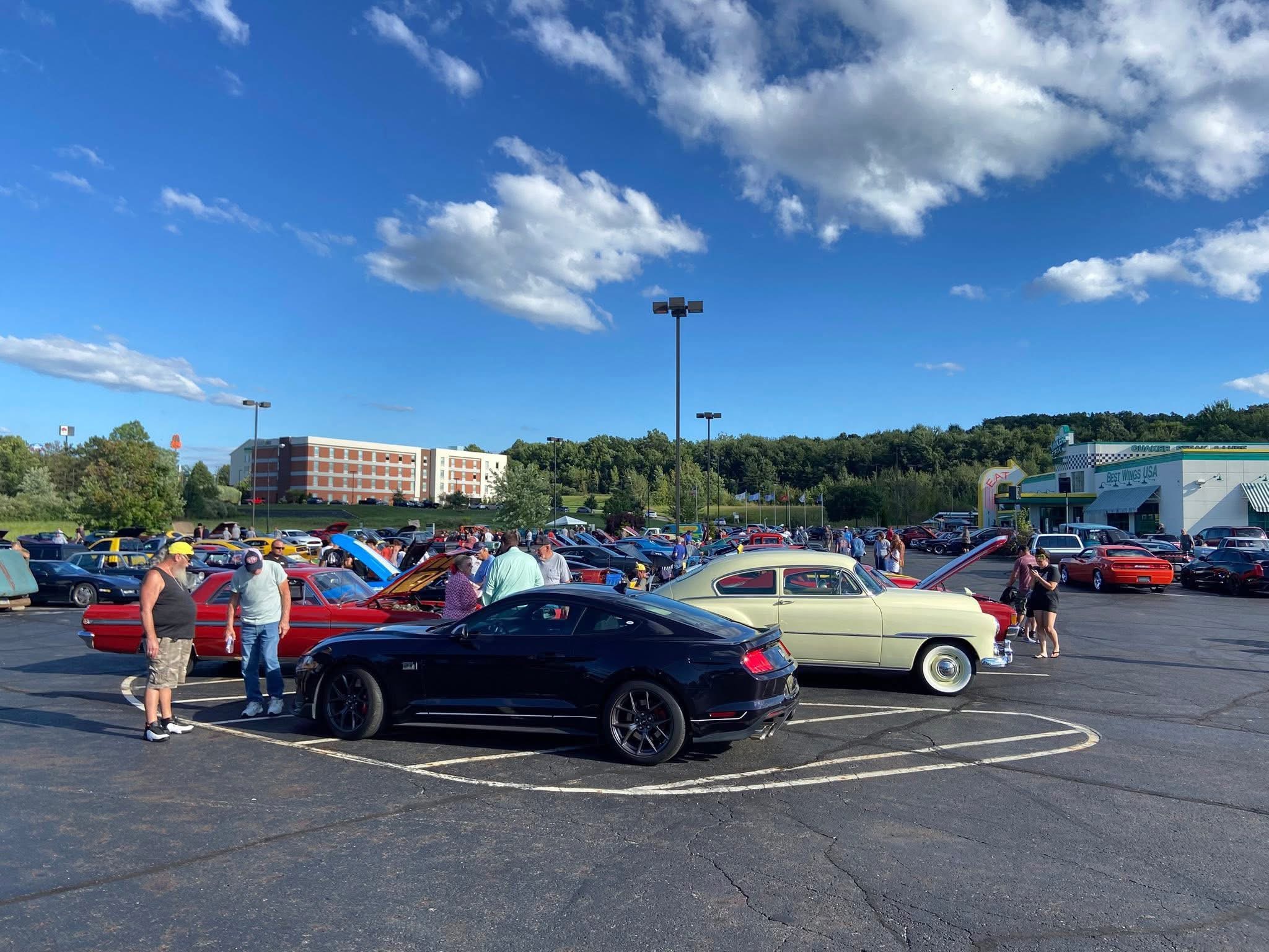 People gather around classic and modern cars in a parking lot on a sunny day with clouds.