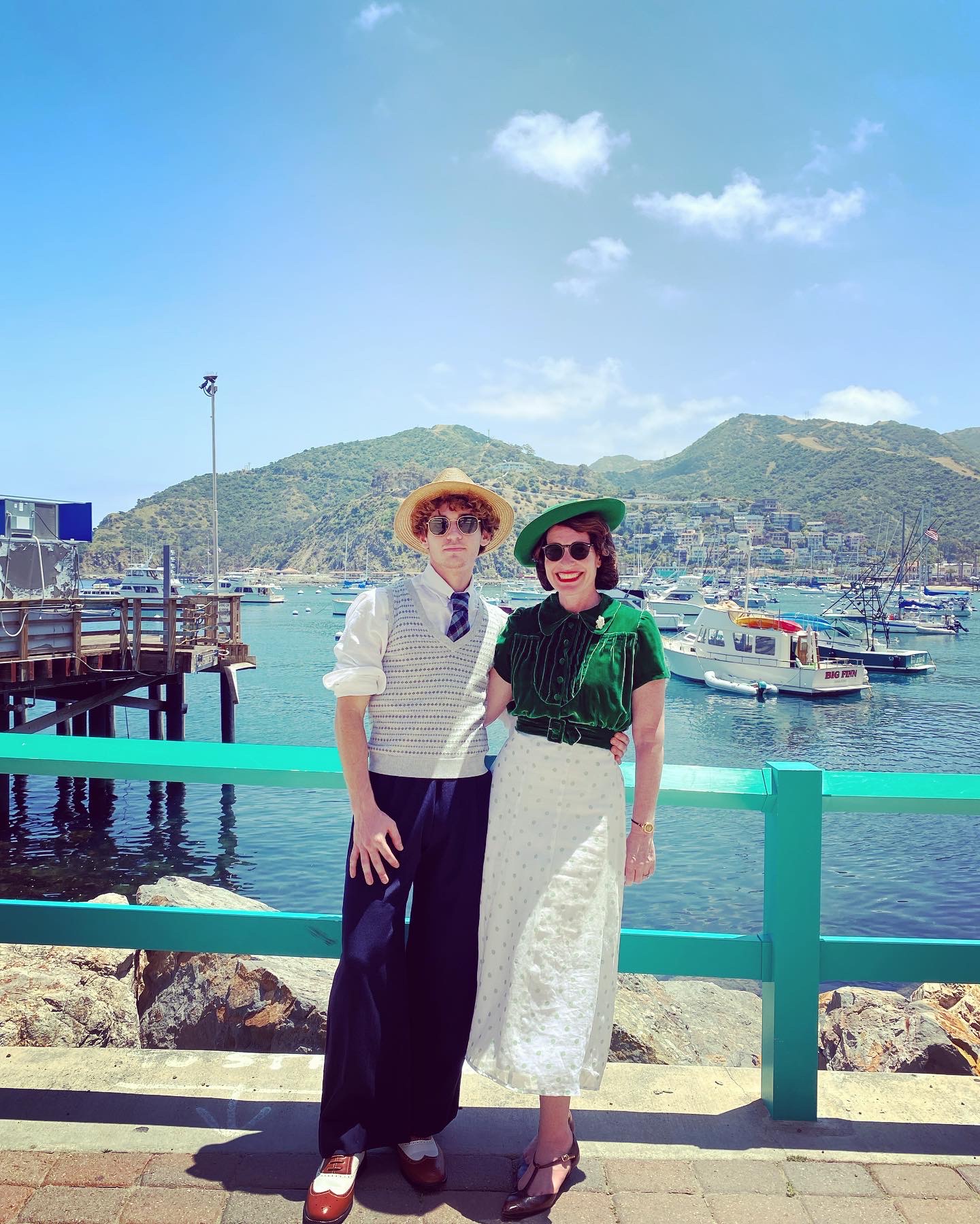 Two women stand by a turquoise railing near a marina with boats and hills in the background, sunny sky with a few clouds.